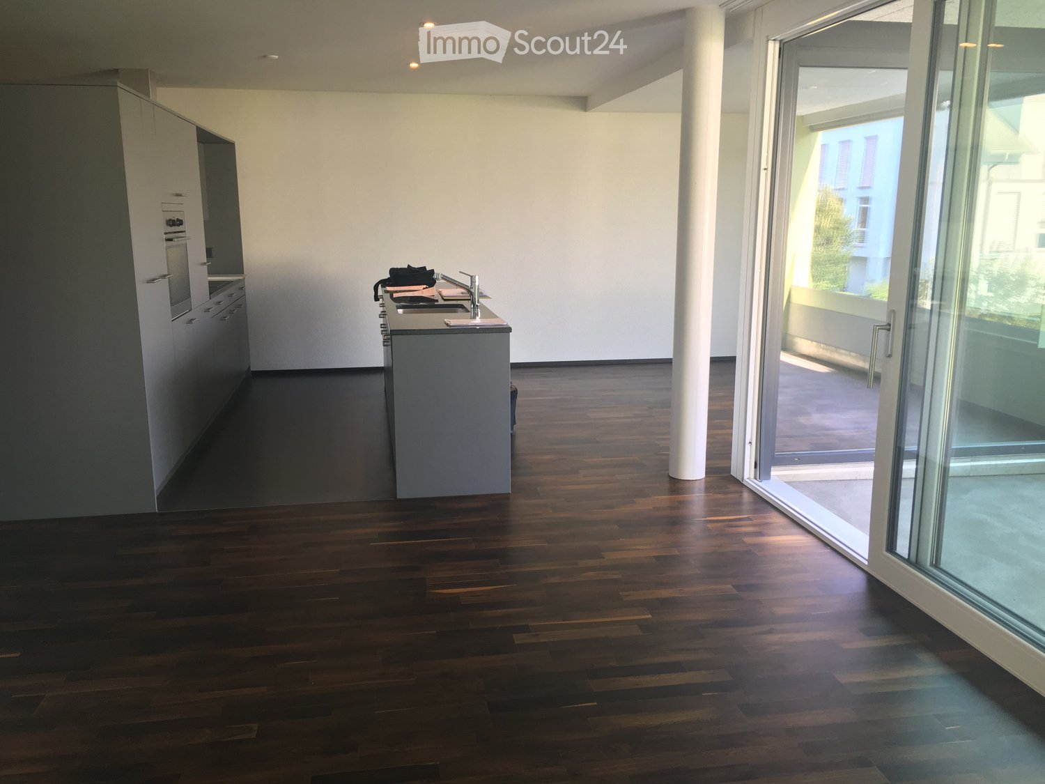 Modern kitchen with dark wooden flooring, white cabinets, and a central island with a sink and faucet. Large glass doors lead to a balcony.