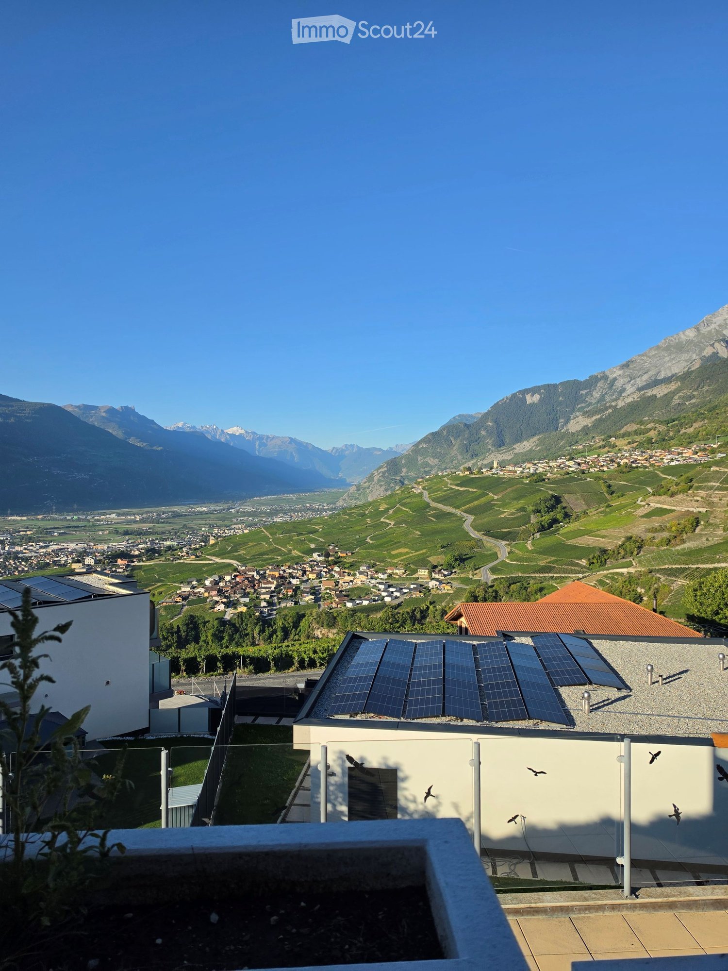 Balcony with solar panels, overlooking a town and mountain landscape.