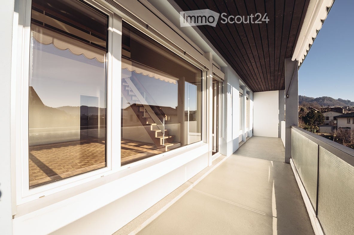Spacious balcony with large windows, wooden flooring, and a view of the surrounding landscape.