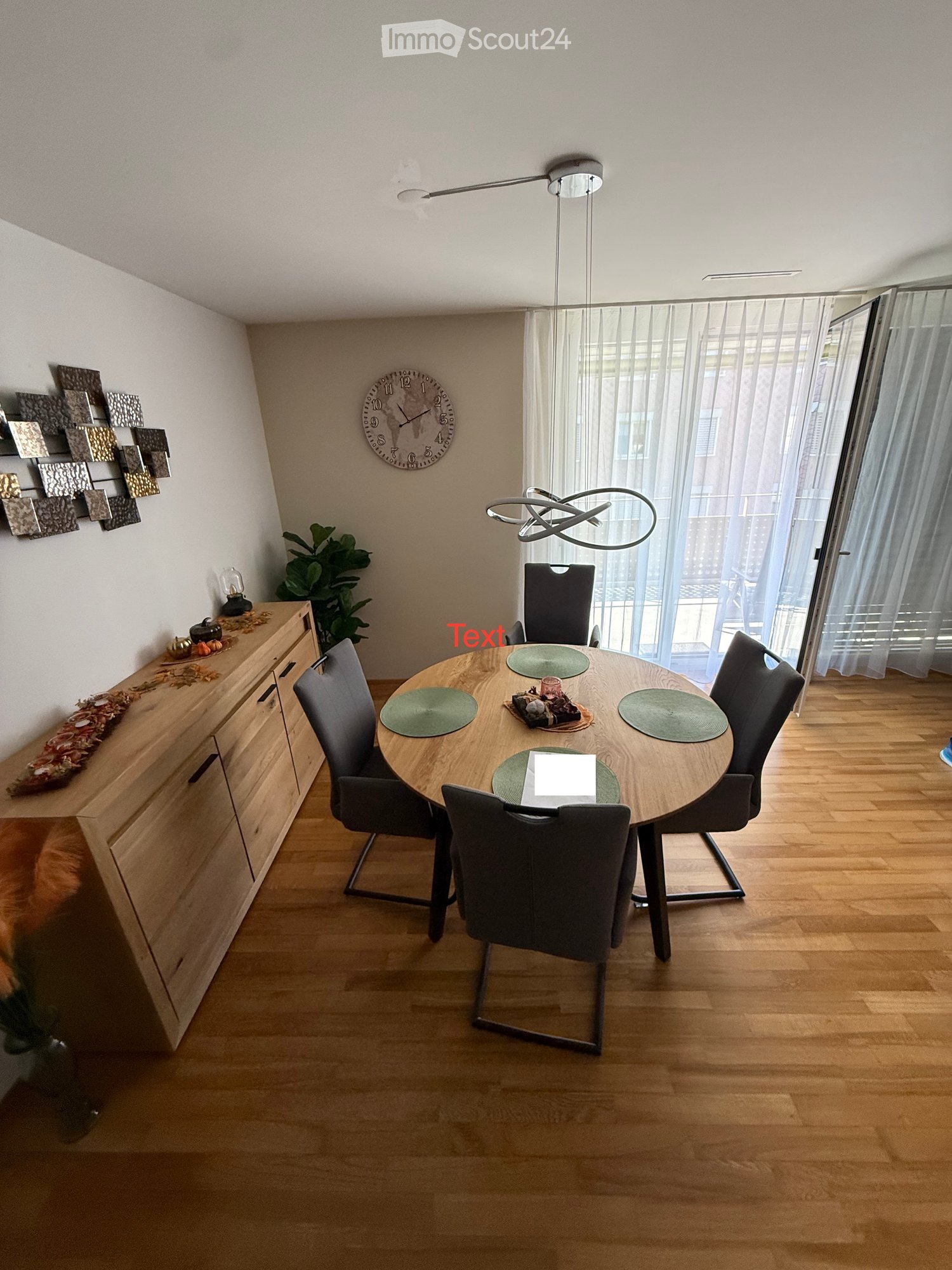 Modern dining room with a round wooden table, four chairs, a hanging lamp, a wall-mounted clock, a wooden cabinet with decorative items, and a potted plant near the window.