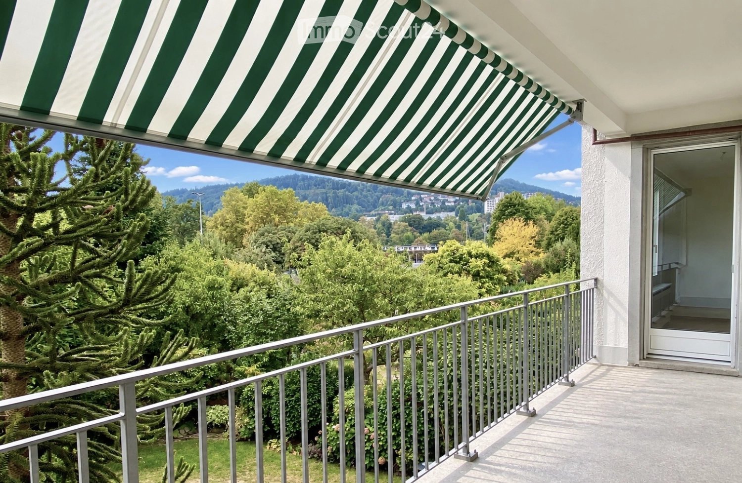 Balcony with metal railing, green and white striped awning, overlooking a lush green landscape with trees and distant hills.