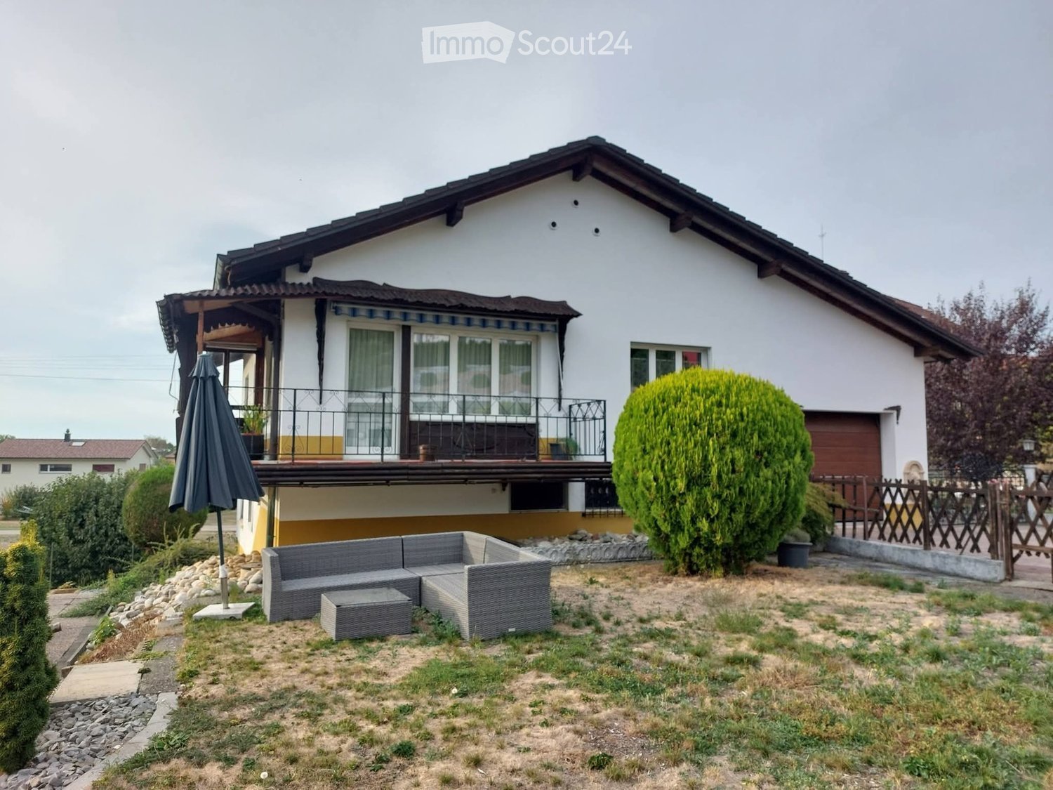 A two-story house with a balcony, surrounded by a garden area featuring a grey outdoor sofa, a small table, and an umbrella. The garden includes a large, well-maintained bush and a wooden fence.