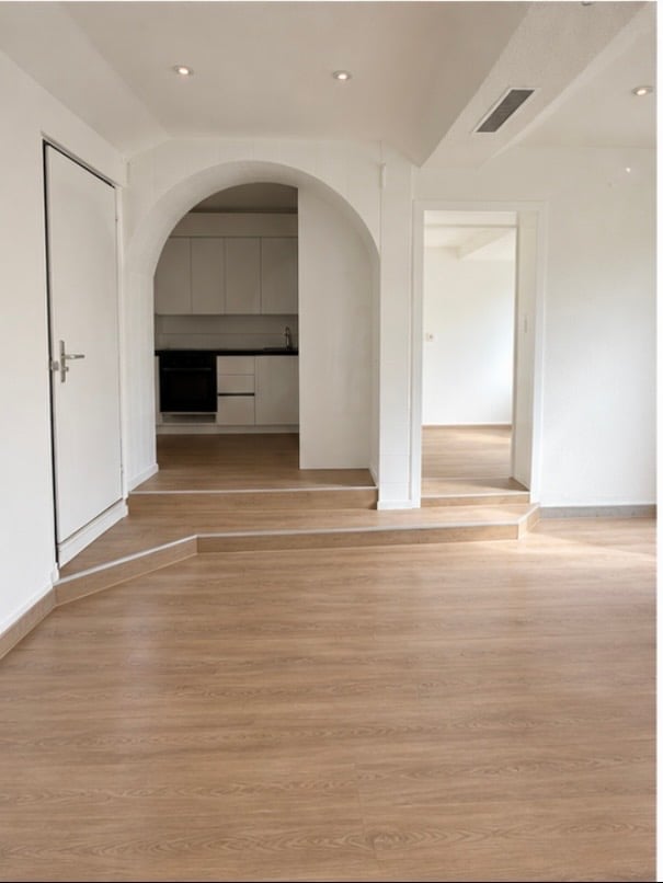 Empty room with light wooden flooring, white walls, and an arched doorway leading to a kitchen area.