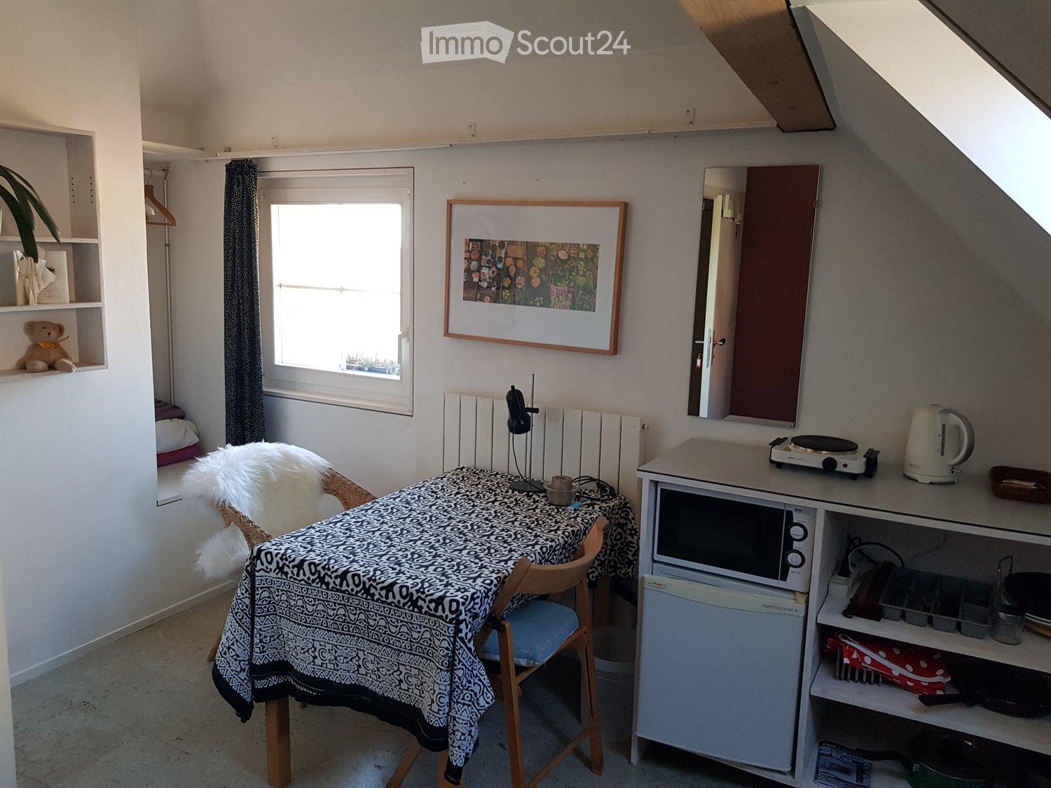 Cozy attic room with a small kitchen area, a dining table with a patterned tablecloth, a chair, shelves, and a window letting in natural light.