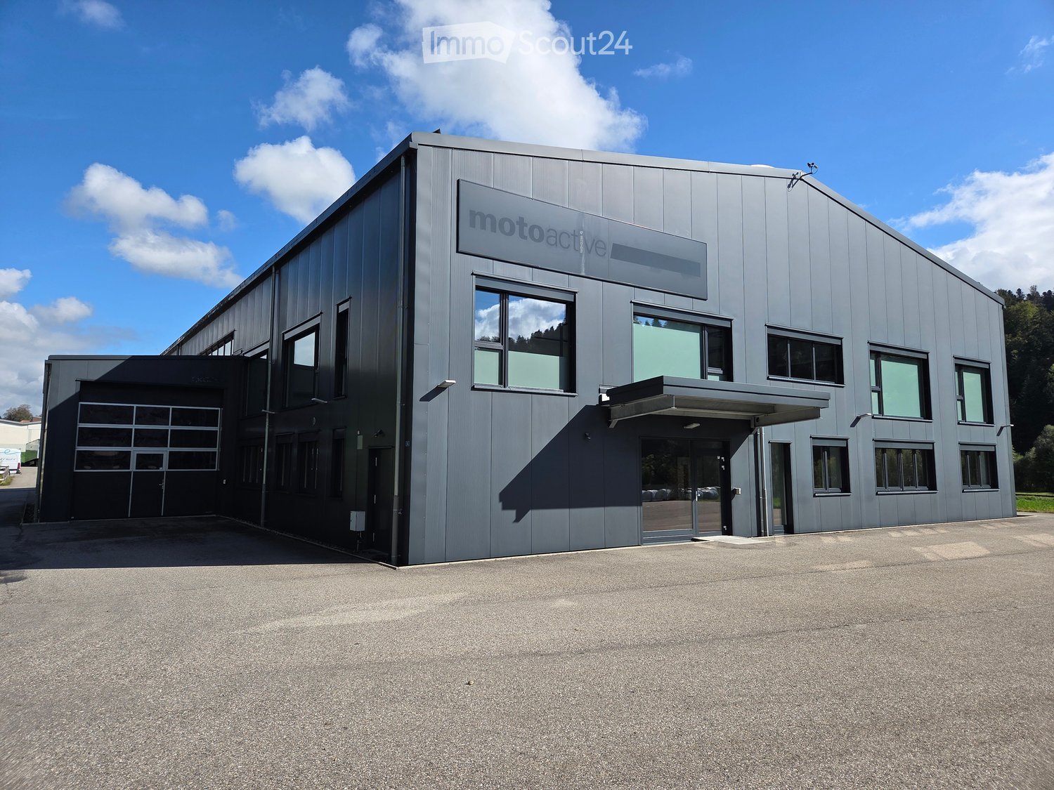 Modern industrial-style building with a grey metal facade, large windows, and a sign reading "motoactive". Features a covered entrance and a spacious parking area.