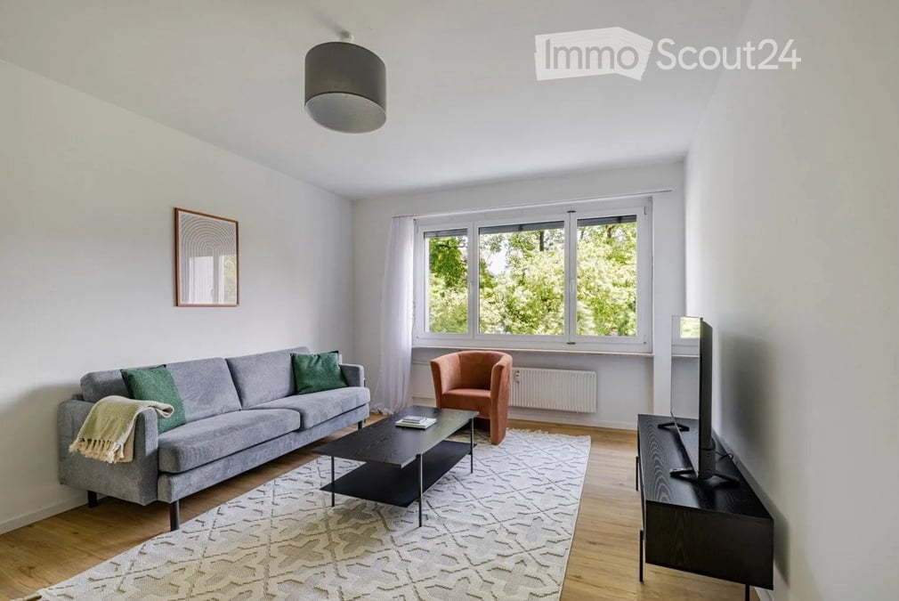 Modern living room with a grey sofa, orange armchair, black coffee table, TV stand, and a large window with white curtains.