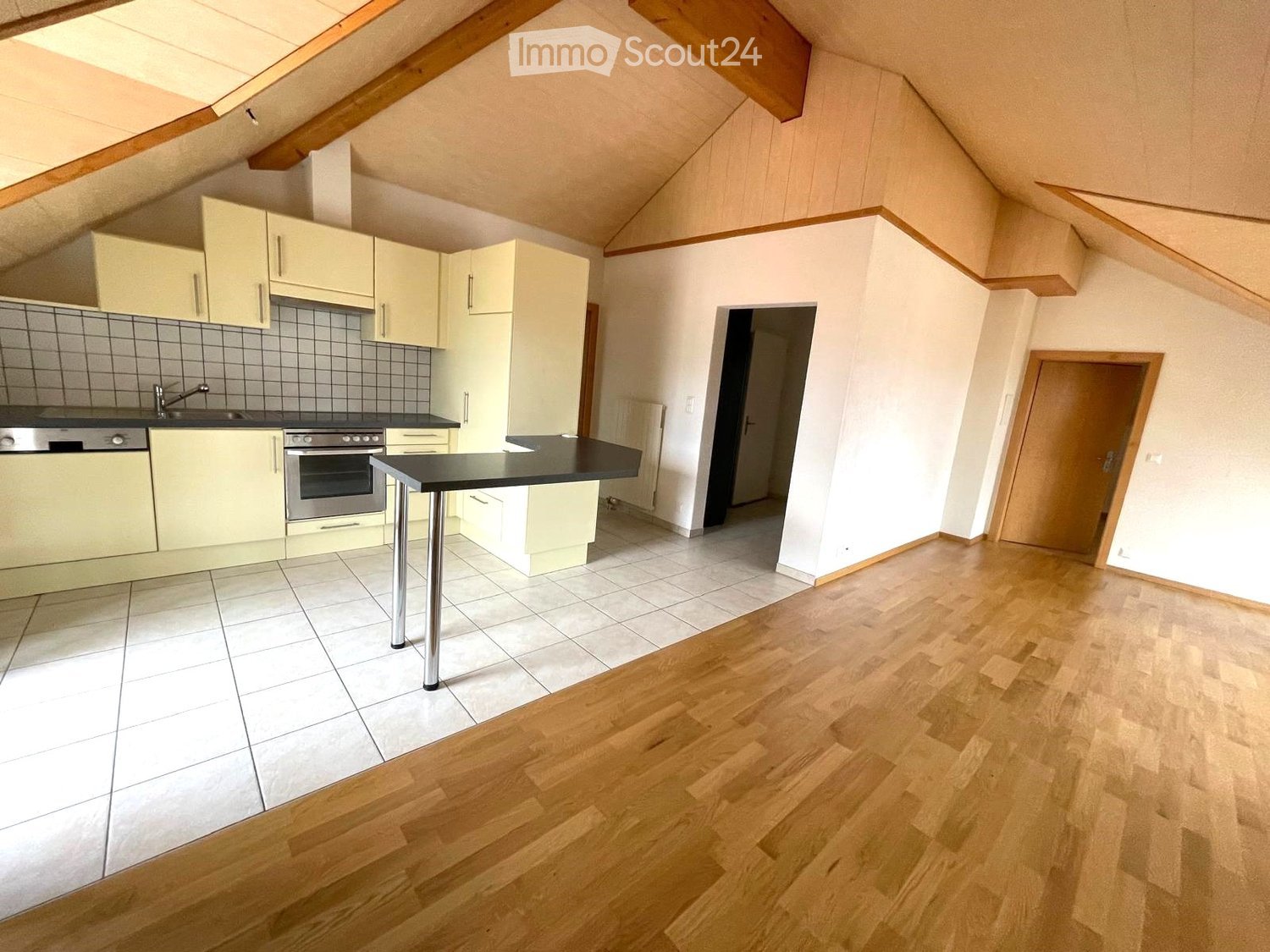 Bright, modern kitchen with light-colored cabinets, stainless steel appliances, and a central island. The space features a mix of tiled and wooden flooring, with a doorway leading to another room.