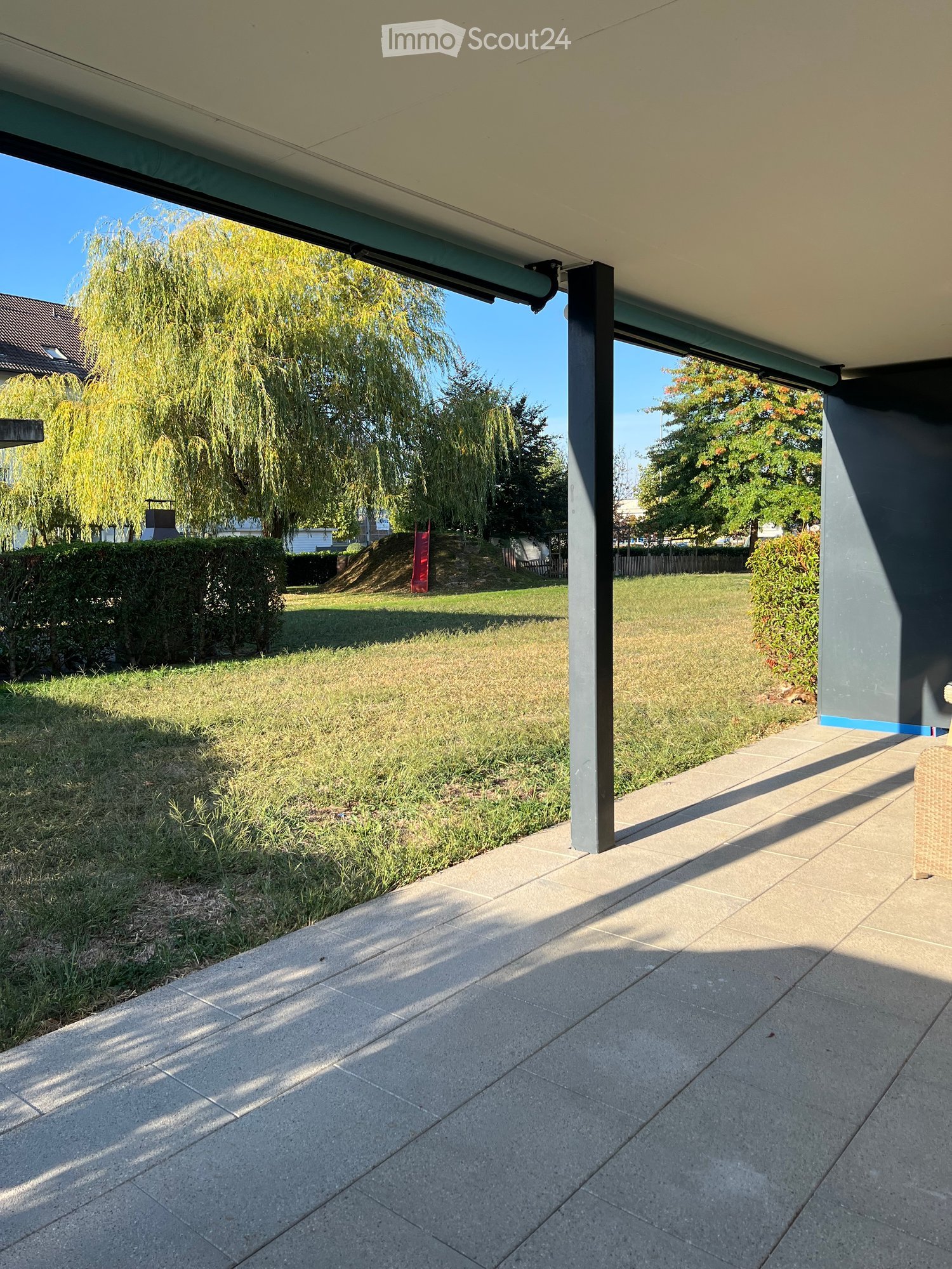 A covered patio area with tiled flooring, surrounded by a grassy lawn, trees, and a hedge. The area is shaded by a modern roof structure with visible support beams.