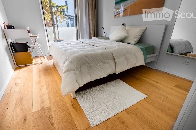 Cozy bedroom with wooden flooring, large window with view, bed with white bedding, nightstand, mirror, small chair, and table.