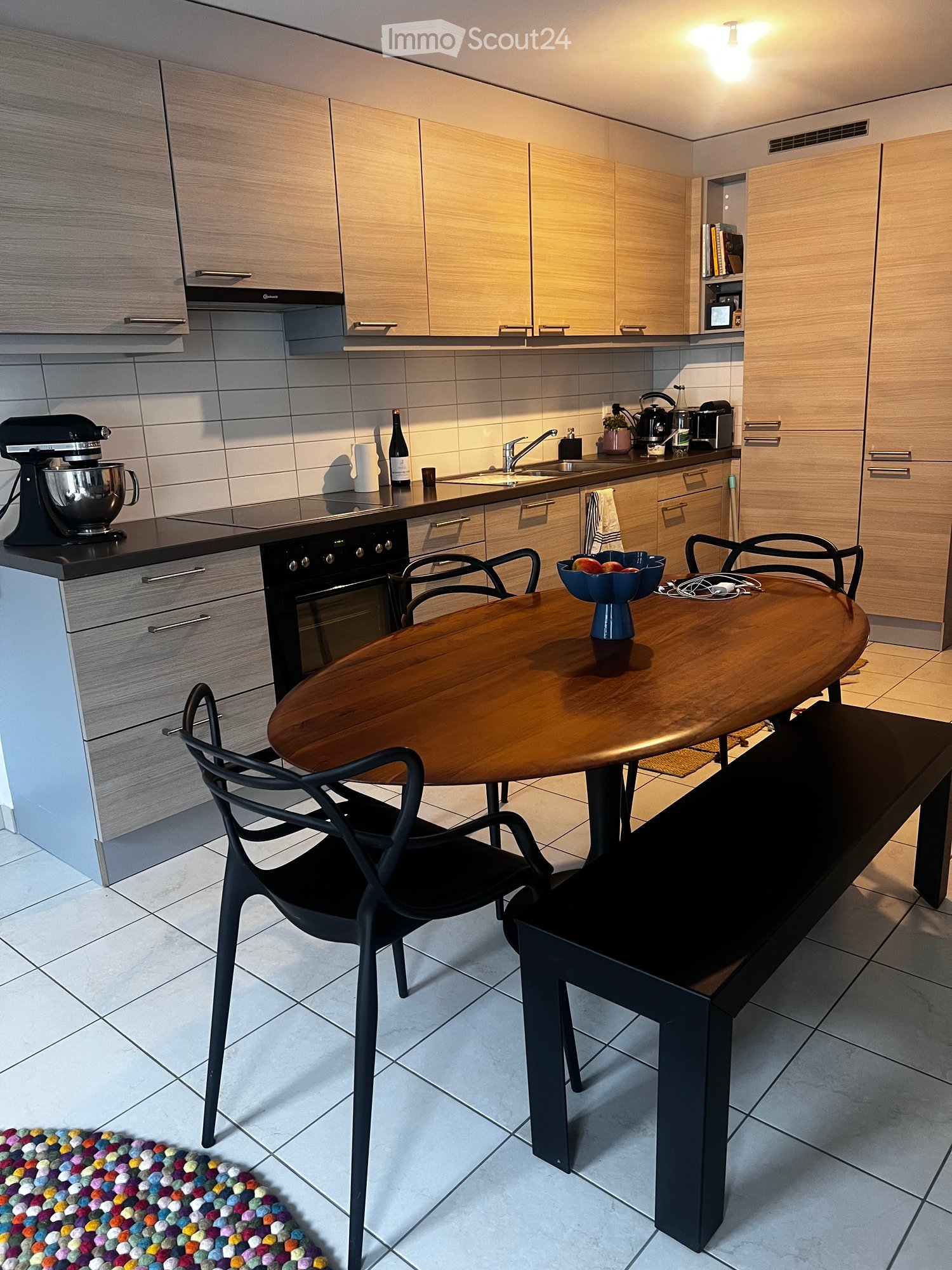 Modern kitchen with light wood cabinets, stainless steel appliances, and a central island with a wooden dining table and black chairs.