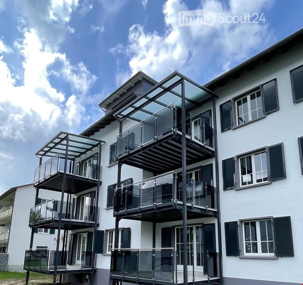 Modern two-story building with white exterior, multiple large windows with shutters, and several glass-enclosed balconies.