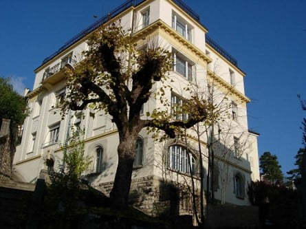 A multi-story building with a balcony on the top floor, surrounded by trees.