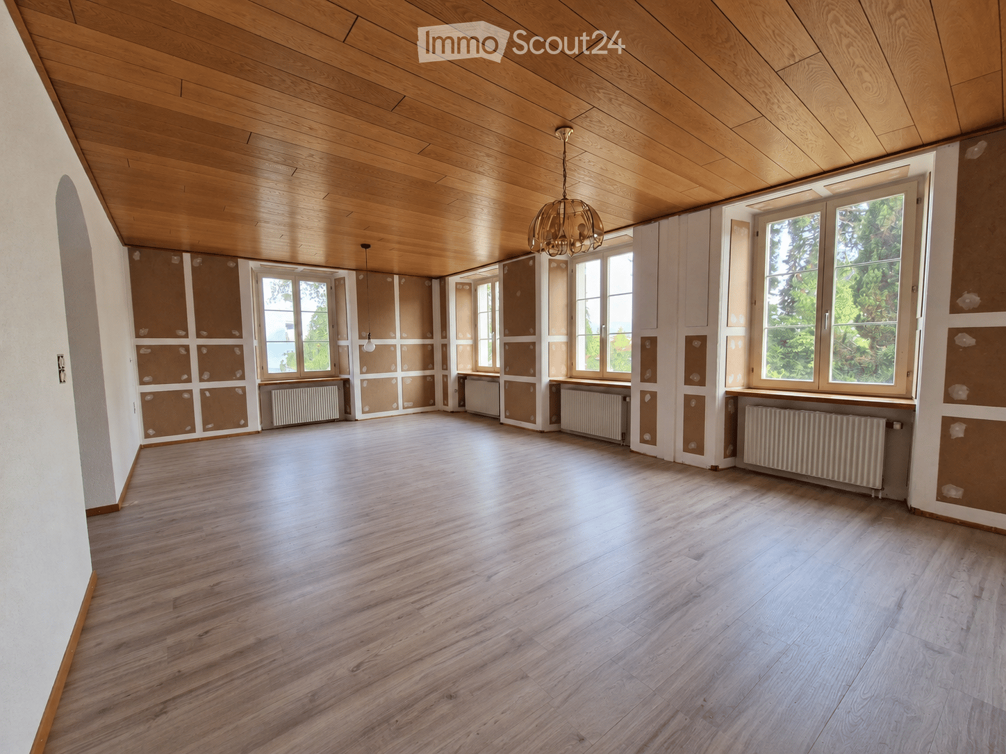 Empty room with wooden flooring and wooden ceiling, large windows with radiators below, and exposed wooden beams on the ceiling.