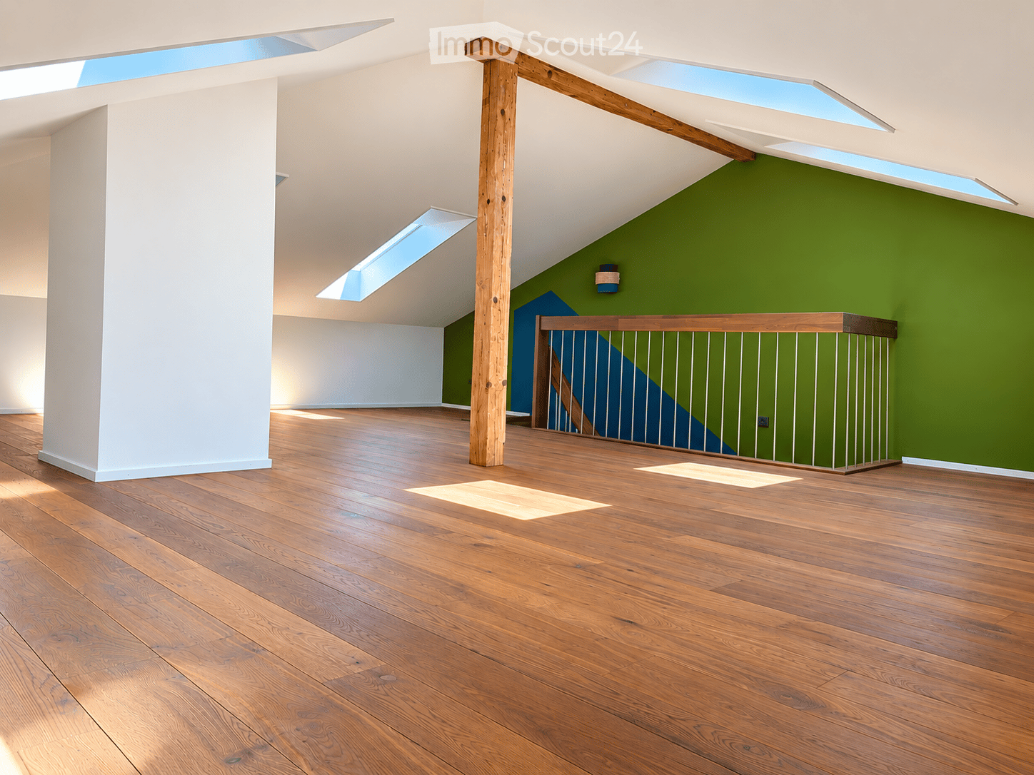 Empty room with wooden flooring, white walls, and skylights allowing natural light. Features a wooden post and a metal railing leading to another area.
