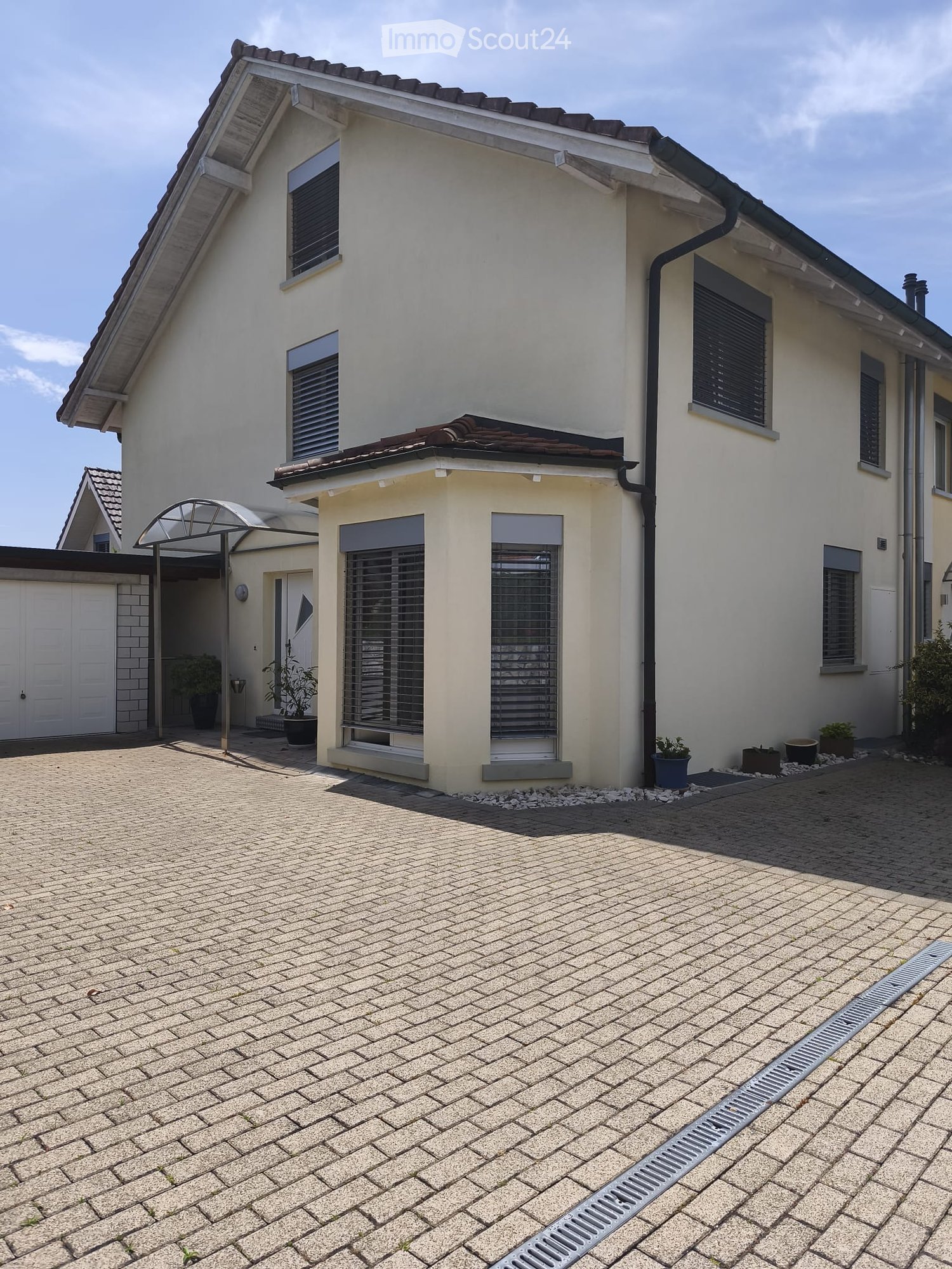 Two-story house with light-colored exterior, multiple windows with shutters, garage, and small garden area with potted plants.