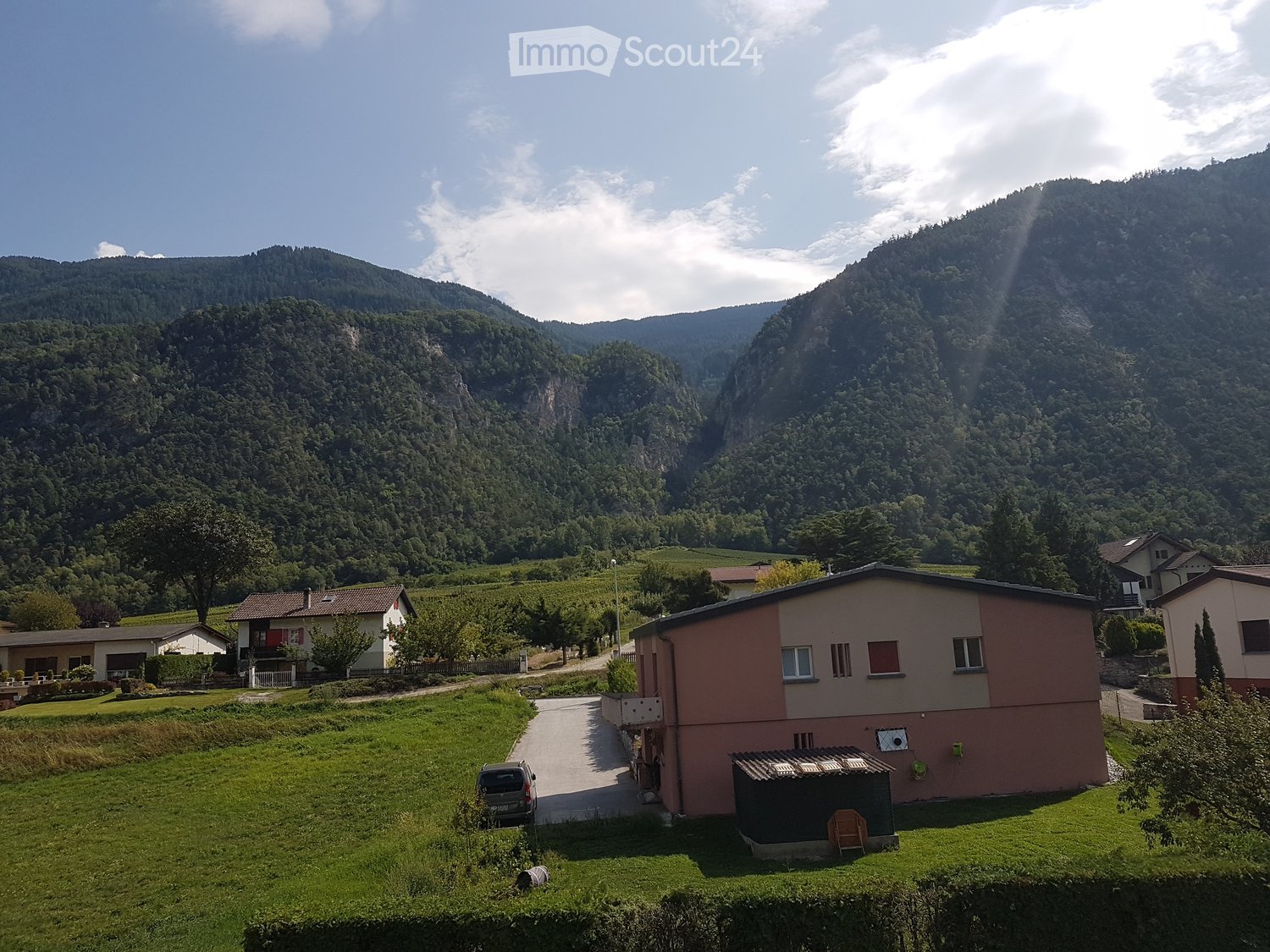 A two-story house with a pink facade and white trim, situated in a lush green valley surrounded by mountains. The house features a driveway, a small shed, and a parked car. The surrounding area includes vineyards and other houses, with a scenic mountain b