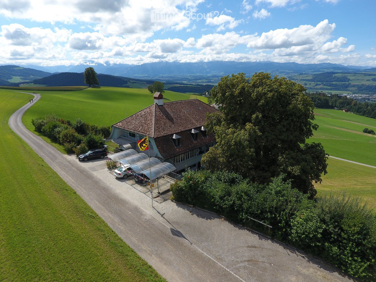 Aerial view of a two-story building with a brown roof, surrounded by green fields and trees, with a parking area and a flagpole.