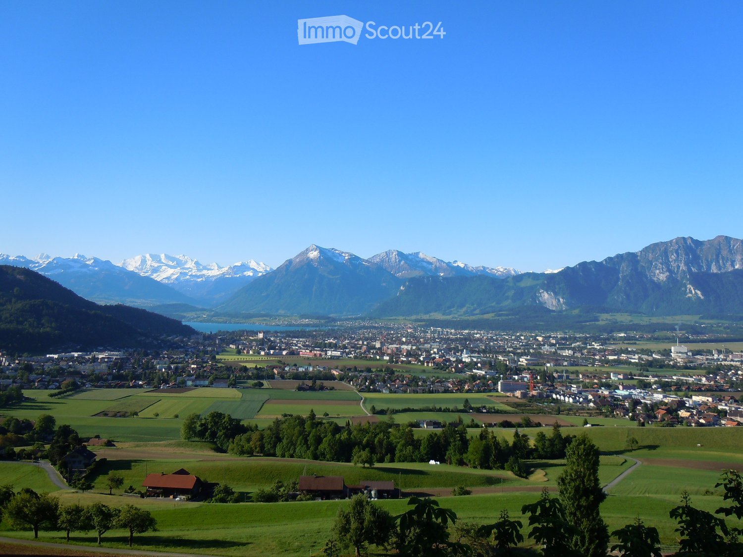 Panoramic view of a town nestled in a valley with surrounding mountains, featuring scattered houses, green fields, and a body of water in the distance.
