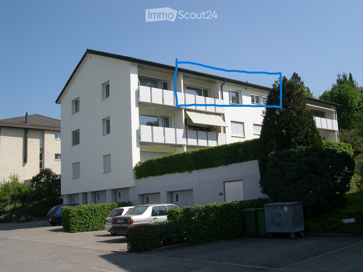 A modern, multi-story residential building with white exterior walls, multiple balconies, and a well-maintained garden area. The building features large windows and a clean, contemporary design.