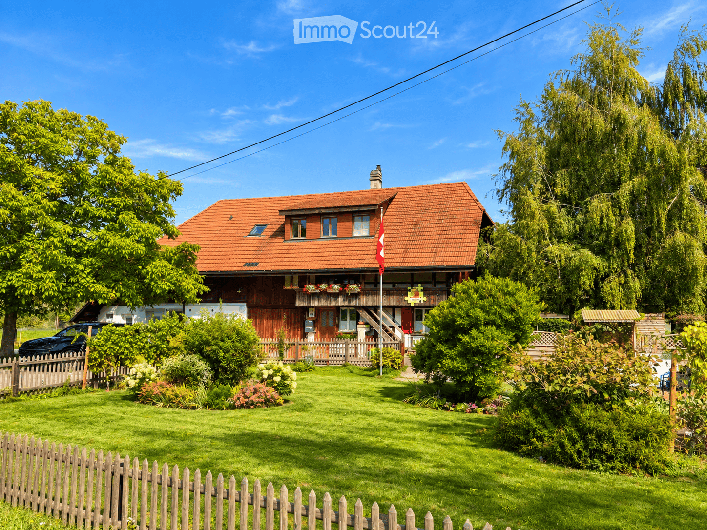 Two-story house with red-tiled roof, wooden balcony, and well-maintained garden with a wooden fence.