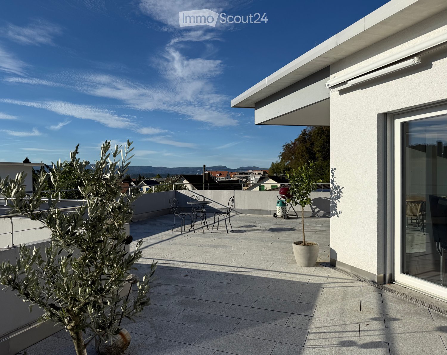 Modern rooftop terrace with tiled floor, small table and chairs, potted plants, and a view of the city and mountains.