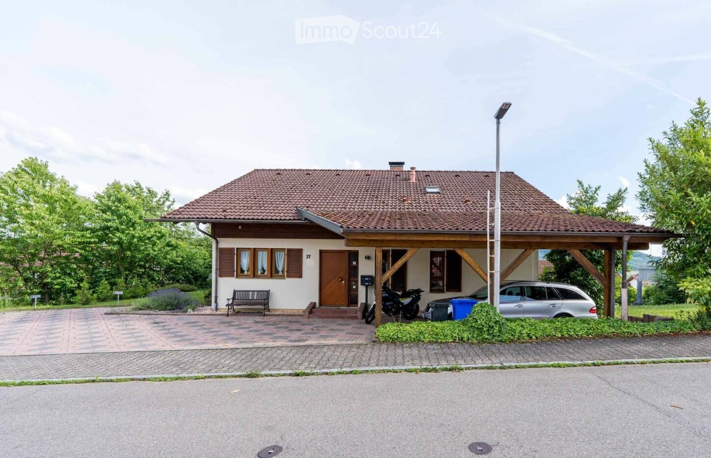 Single-story house with a tiled roof, wooden entrance, and covered porch area. The house has multiple windows with shutters, a bench near the entrance, and a driveway with parked vehicles including a car and motorcycles.