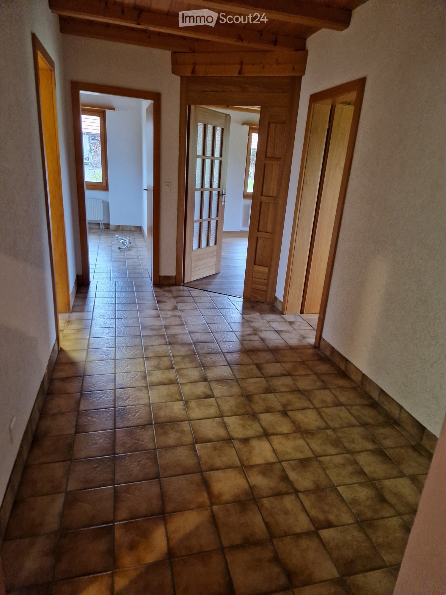 A spacious hallway with tiled flooring, wooden doors, and natural light from windows at the end. The walls are painted in a neutral tone, and the ceiling features exposed wooden beams.