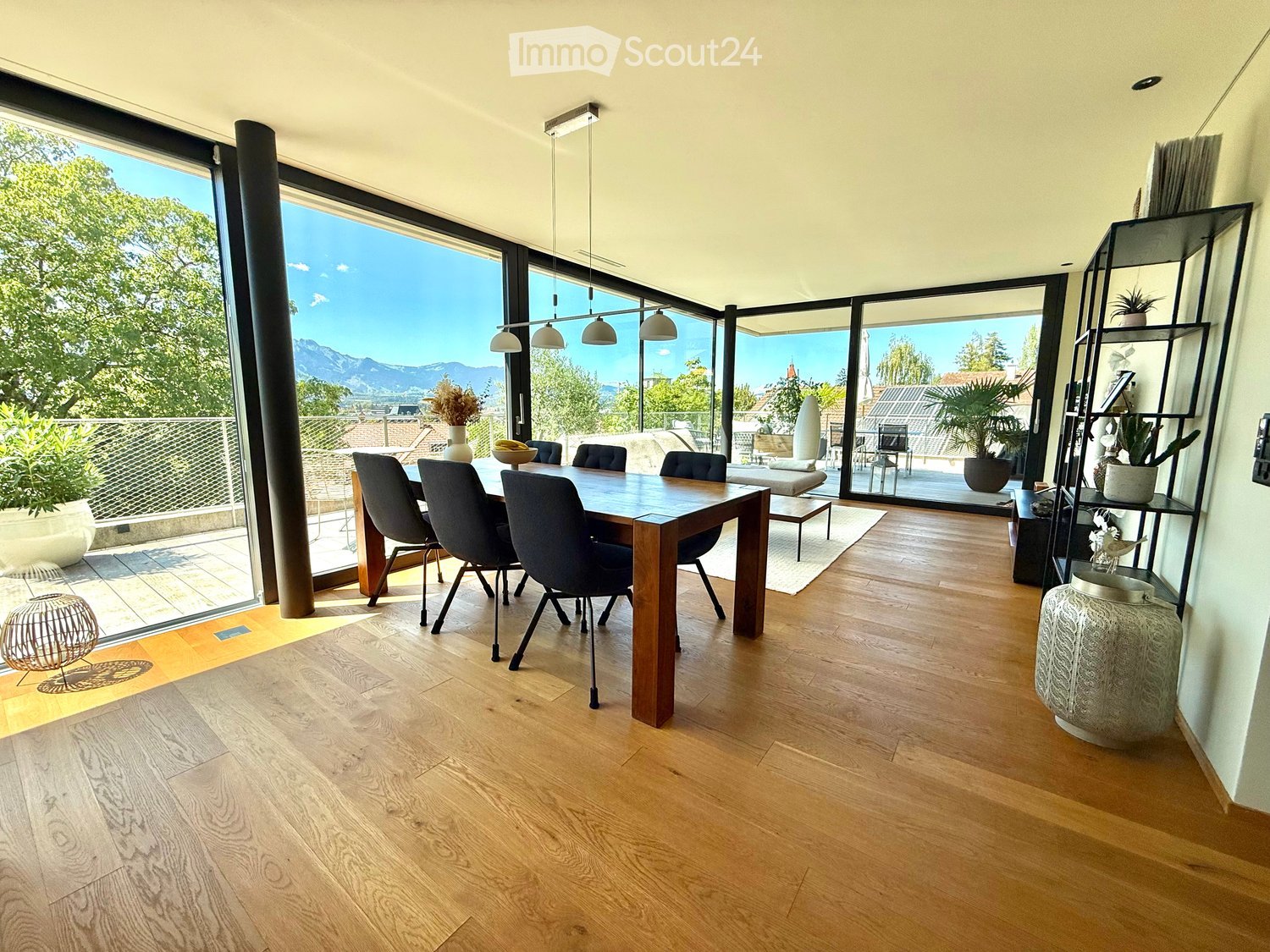 Bright dining area with wooden floor, large windows offering a scenic mountain view, dining table with chairs, decorative shelf, and potted plants.