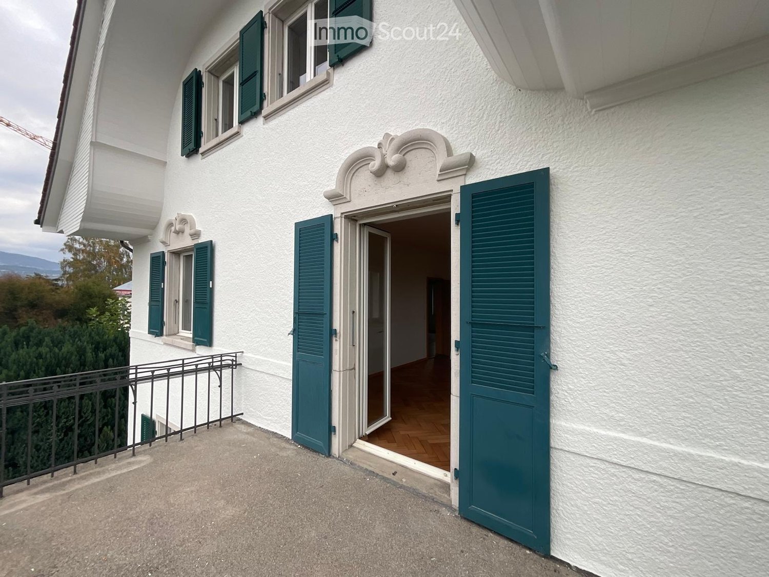 Balcony with metal railing, green shutters, and a view of the house entrance and surrounding greenery.