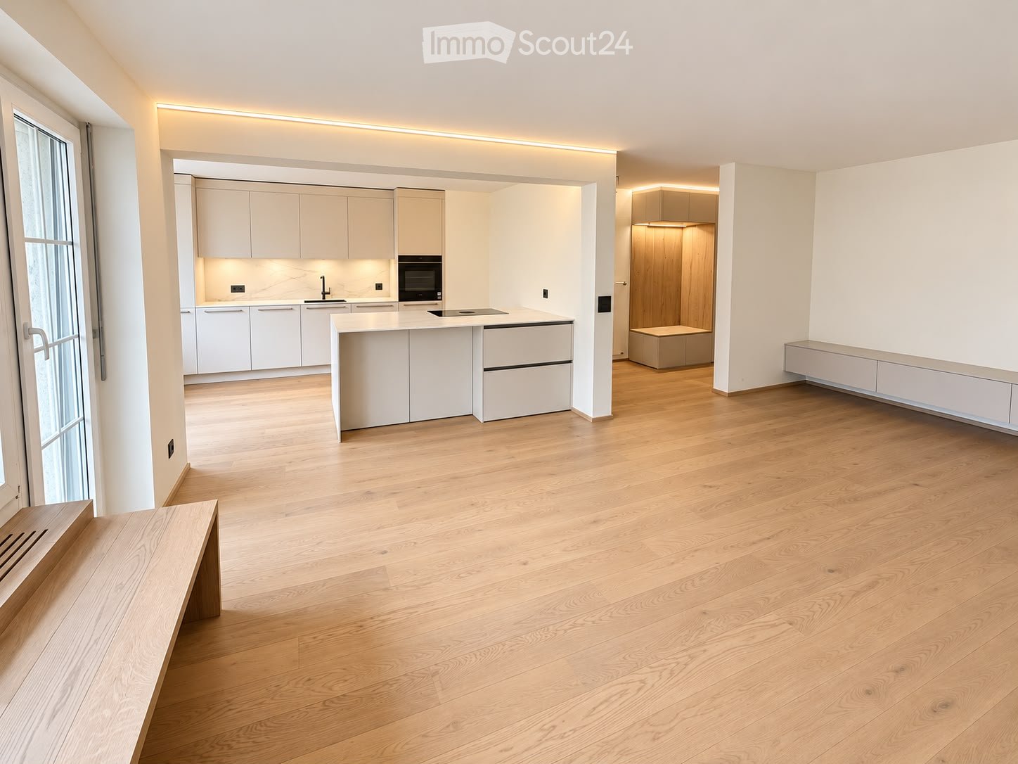 Open-plan space with light wooden flooring, white cabinetry, and built-in appliances in a modern kitchen area. The room is empty, with no furniture or personal items.