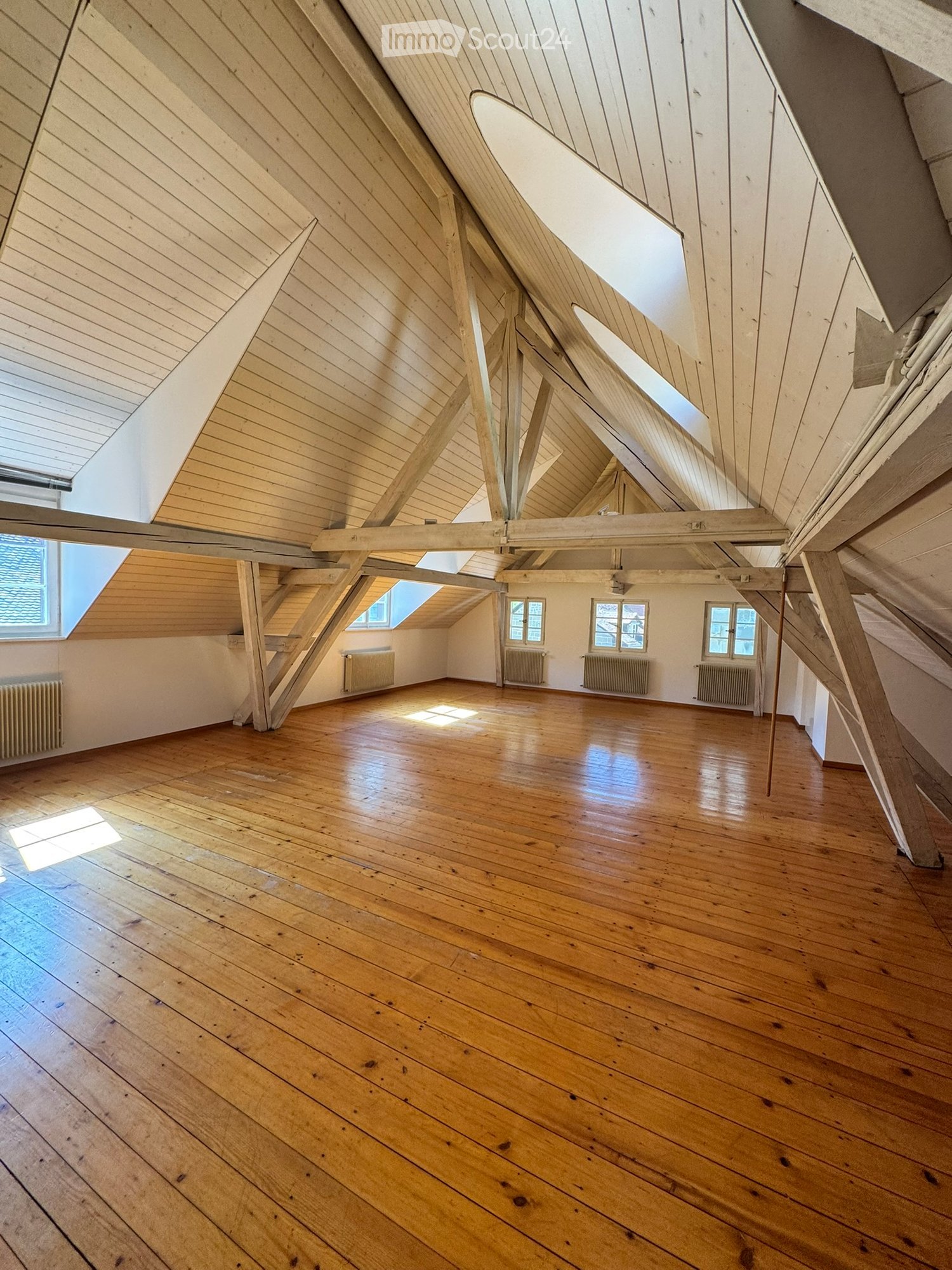 An empty attic space with wooden floors, exposed wooden beams, and skylights allowing natural light to enter. The room features white walls and radiators along the walls.
