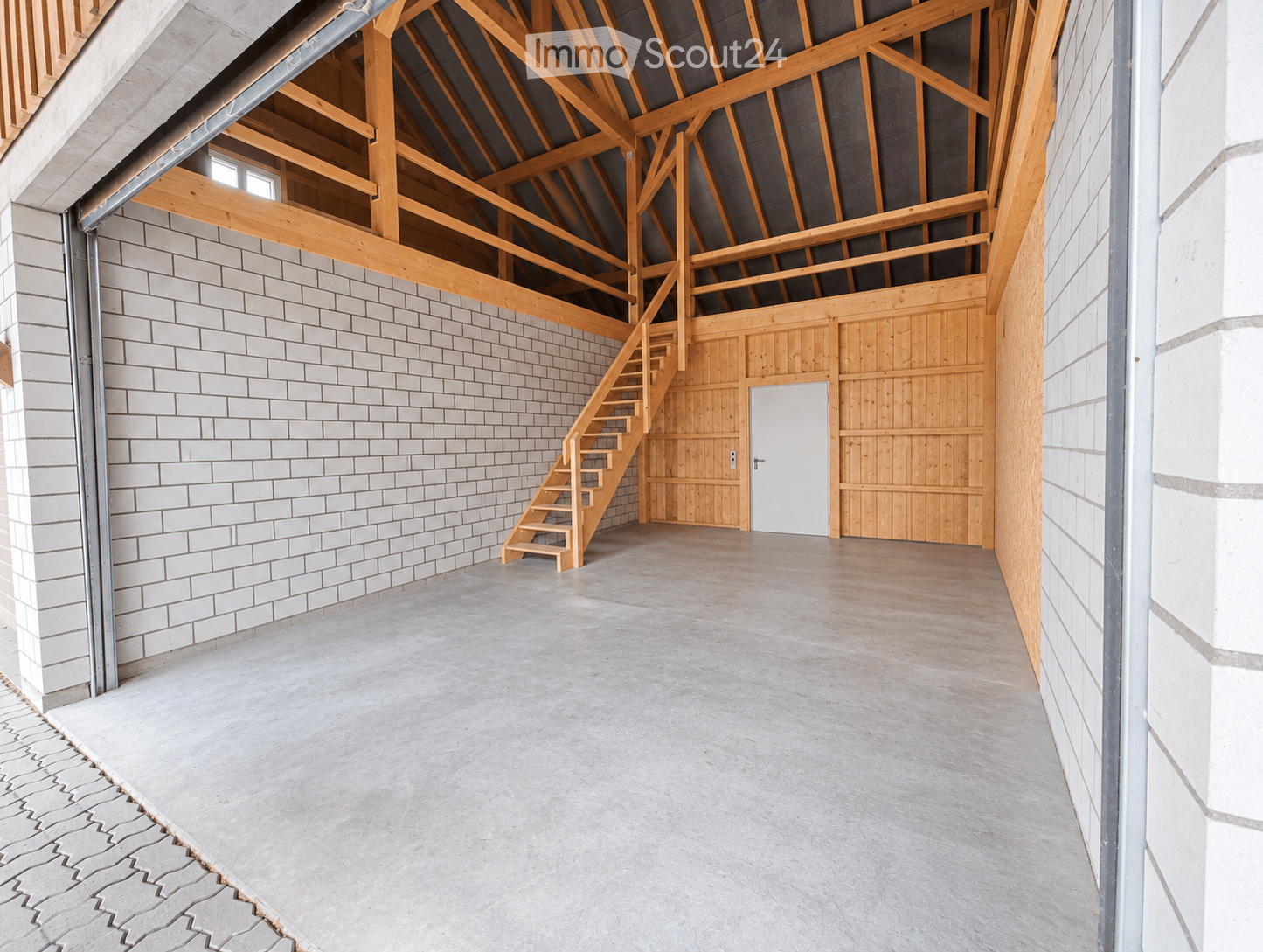 An empty, spacious room with exposed wooden beams and a high ceiling, featuring a staircase leading to an upper level, white brick walls, and a concrete floor.