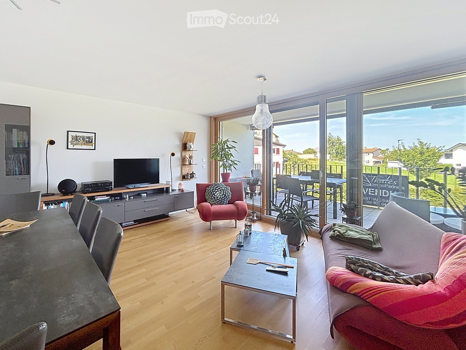Modern living room with wooden flooring, large windows offering a view, a sofa, armchair, coffee table, dining table, TV, bookshelf, and potted plants.