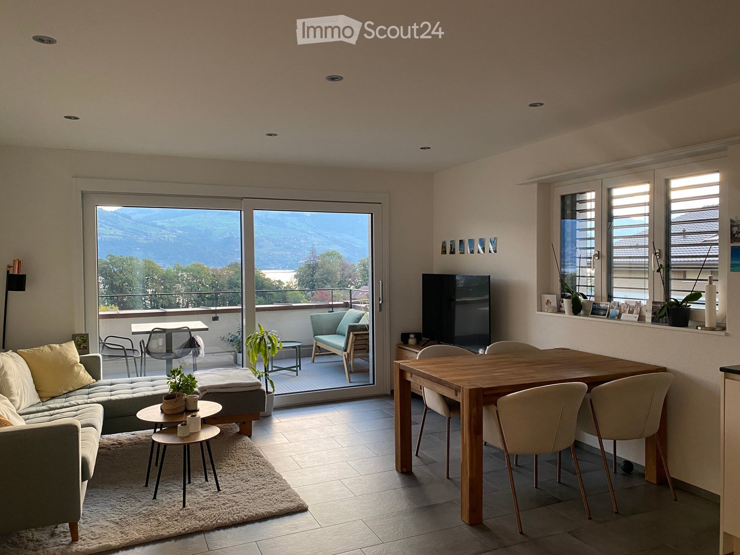 Modern living and dining area with a view, featuring a grey sectional sofa, coffee table, dining table with chairs, and a television.