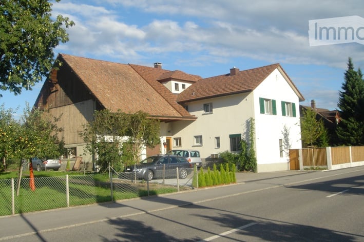 Two-story house with a red-tiled roof, white walls, green shutters, and a fenced front yard with parked cars.