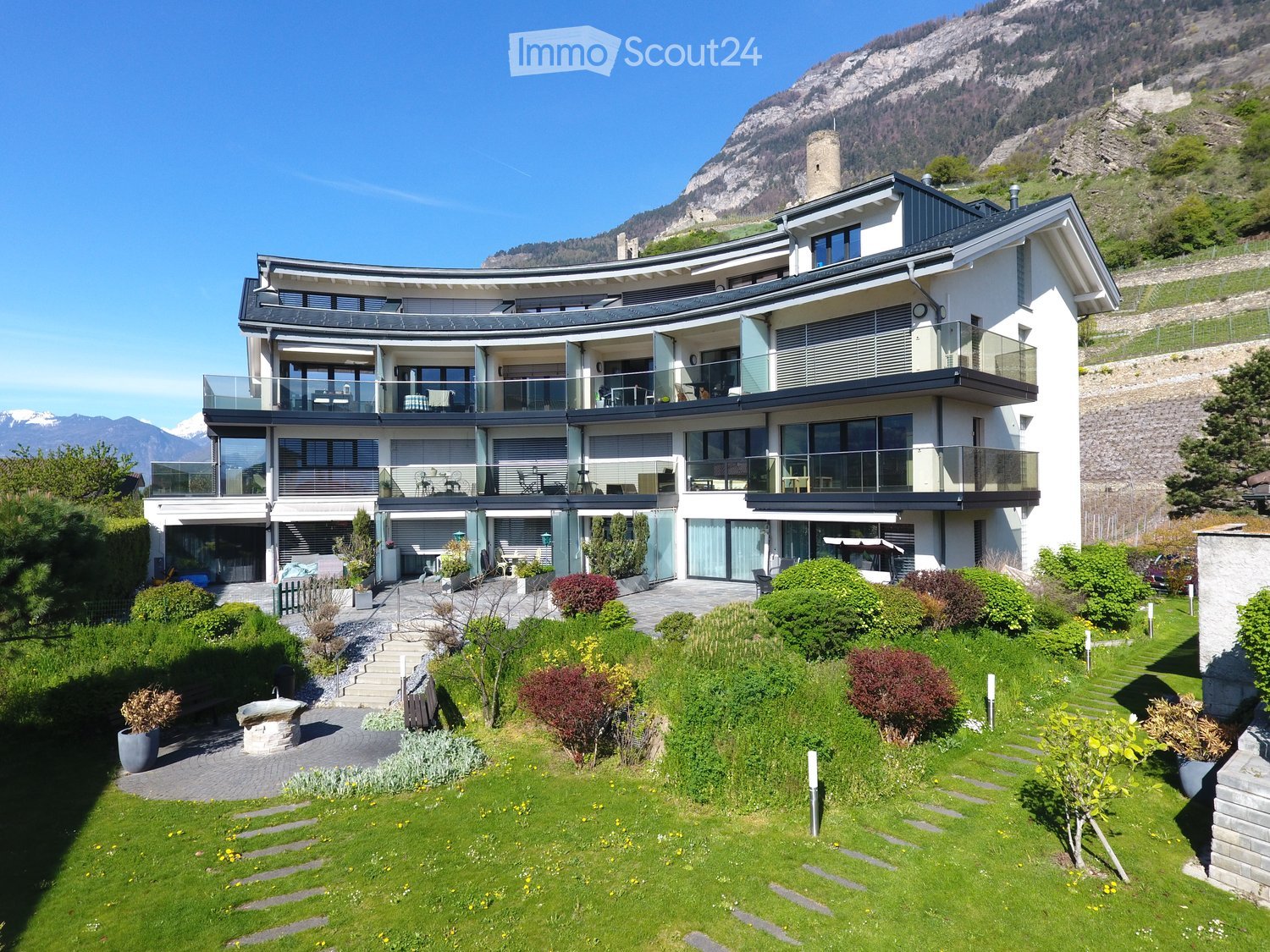 Modern two-story house with large windows and balconies, surrounded by a well-maintained garden with pathways and greenery.