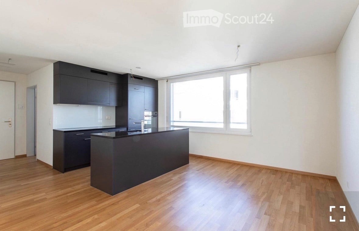 Modern kitchen with dark cabinets, a central island, and a large window providing natural light. The floor is made of light wood.