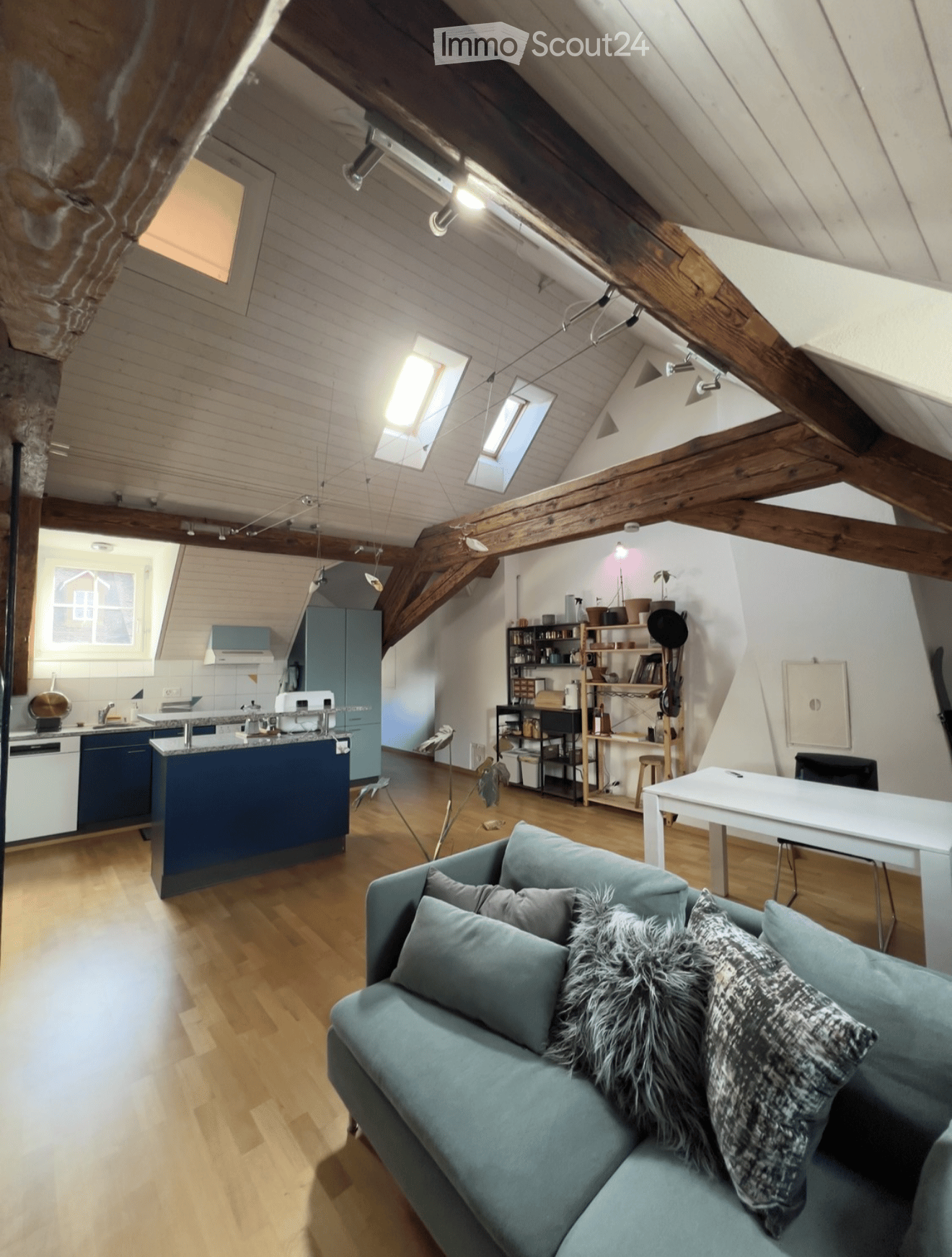Open-plan living and kitchen area with exposed wooden beams, modern kitchen, sofa, dining table, and shelves.