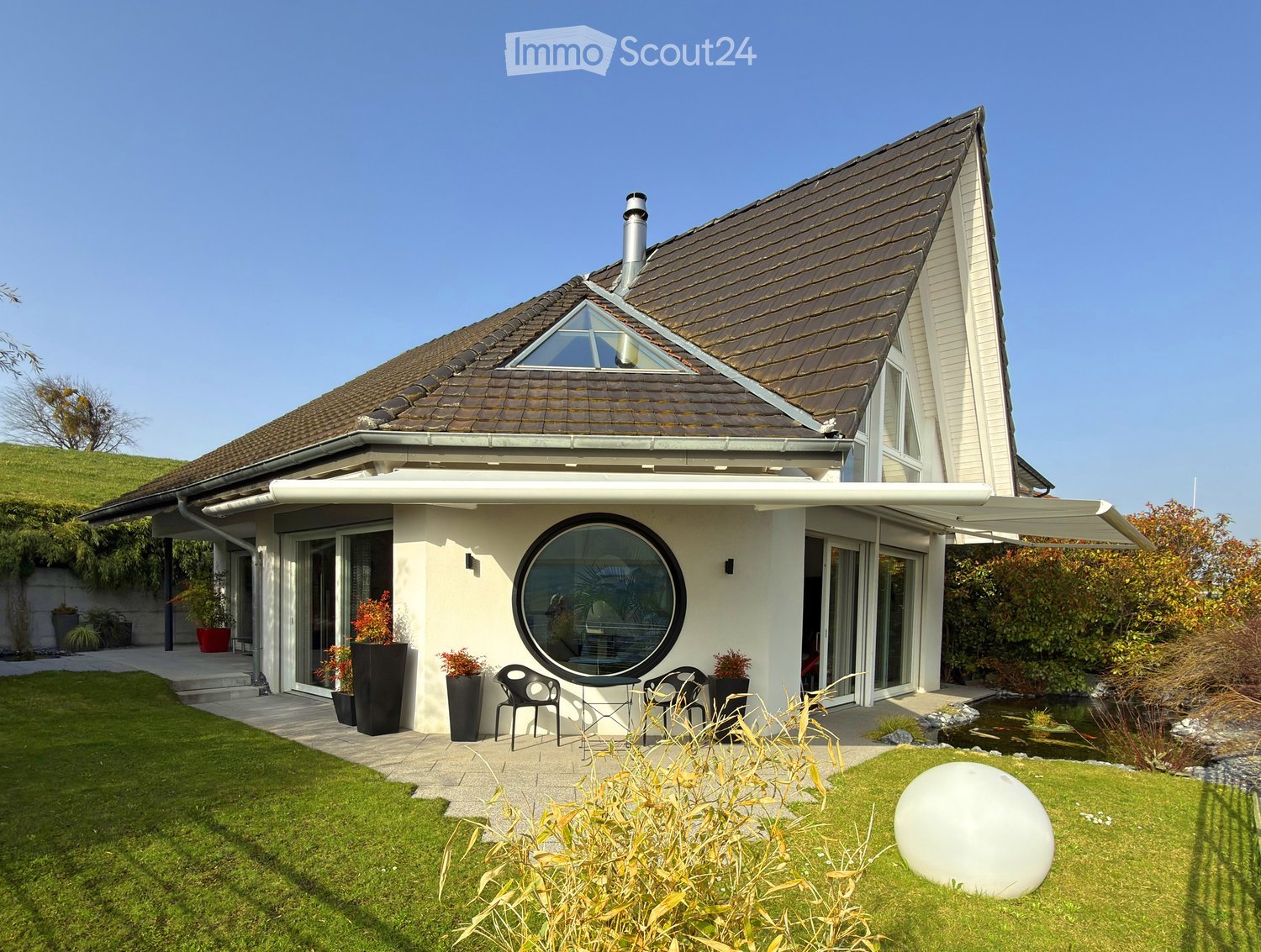 Modern two-story house with a brown tiled roof, large windows, and a spacious garden featuring a pond, patio seating, and decorative plants.