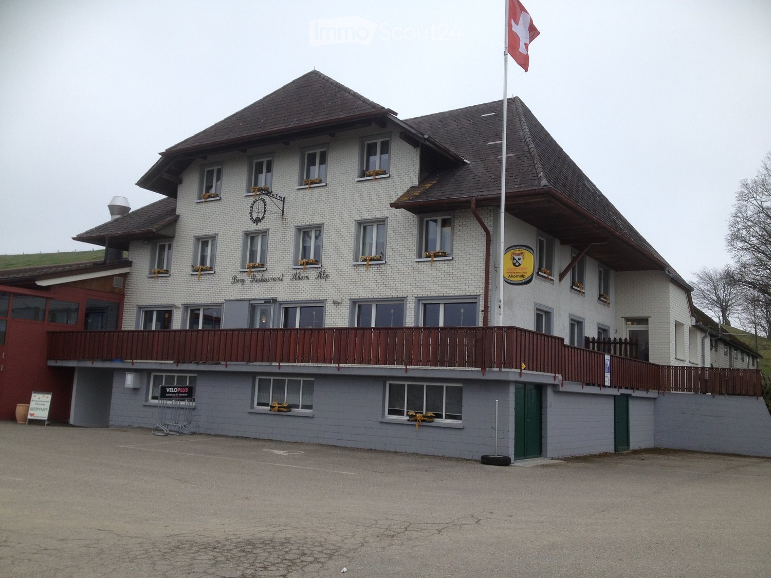 Two-story building with white brick facade, multiple windows, and a balcony with wooden railing. A Swiss flag is displayed on a flagpole, and there is signage for a restaurant named "Berg Restaurant Ahorn Alp." The building has a gray lower level and a br