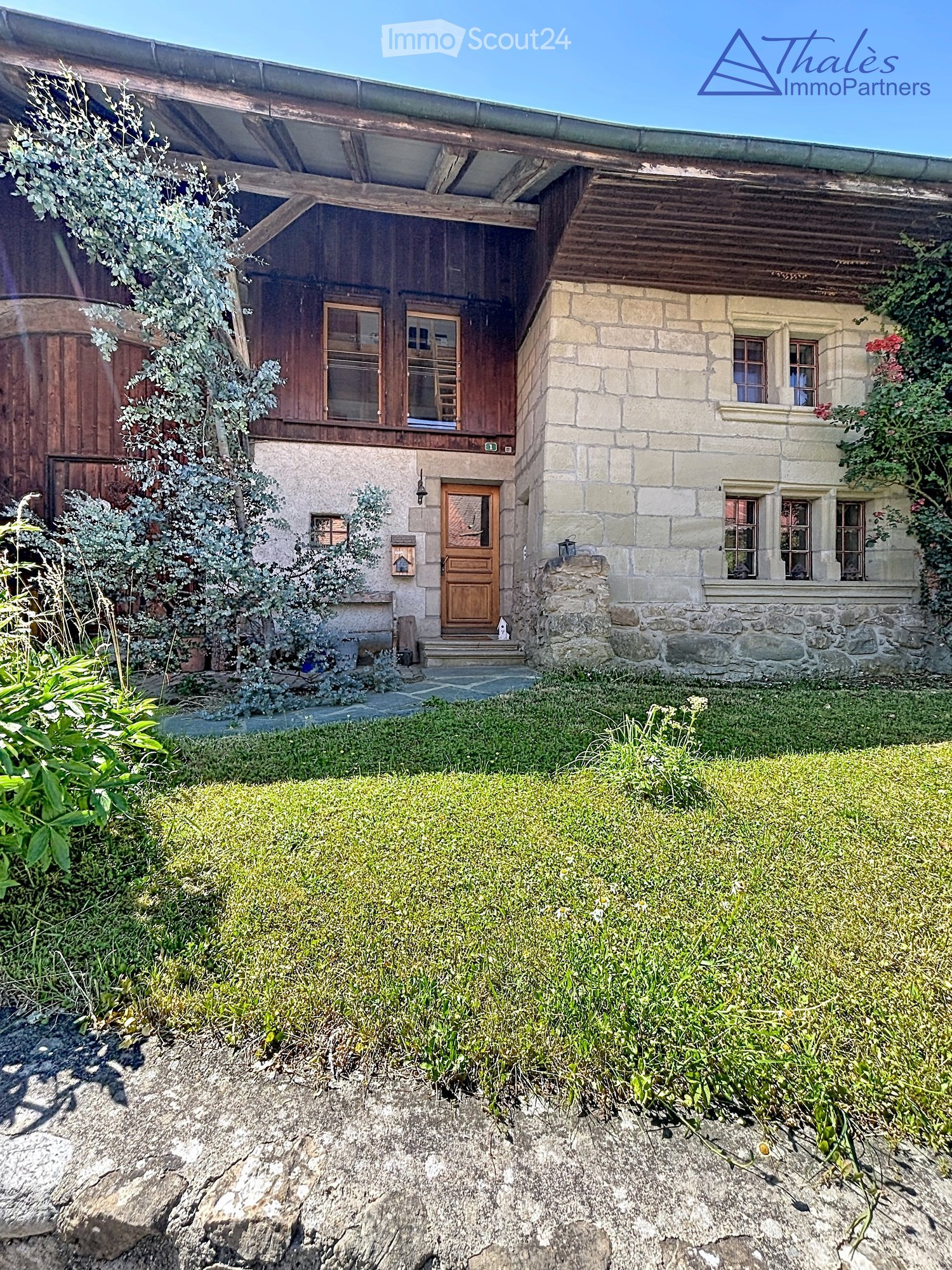 Two-story house with stone and wooden exterior, multiple windows, and a wooden front door. The house is surrounded by a well-maintained lawn and climbing plants.
