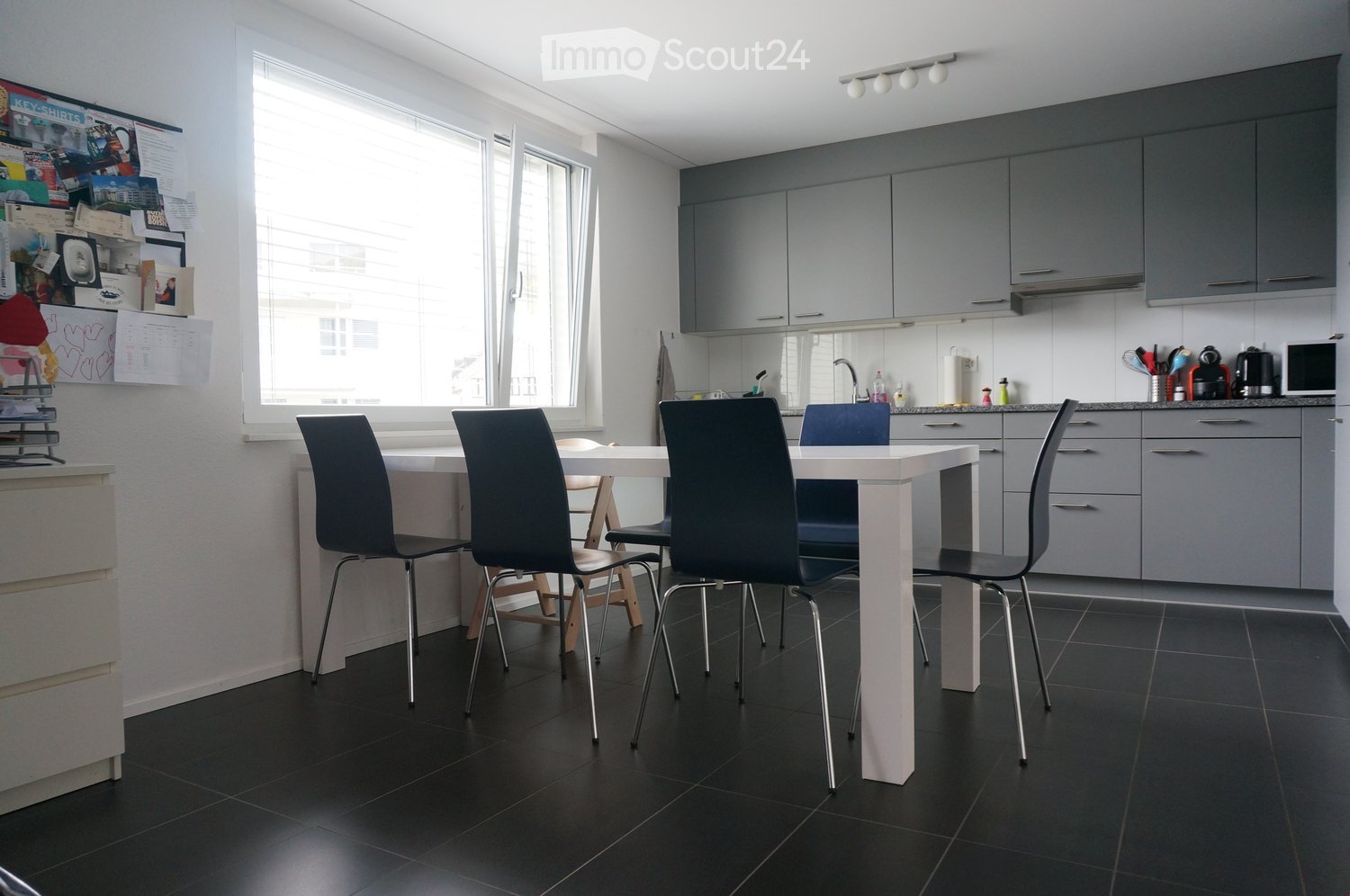 Modern kitchen with grey cabinets, white dining table, four black chairs, and various appliances including a microwave and a faucet.