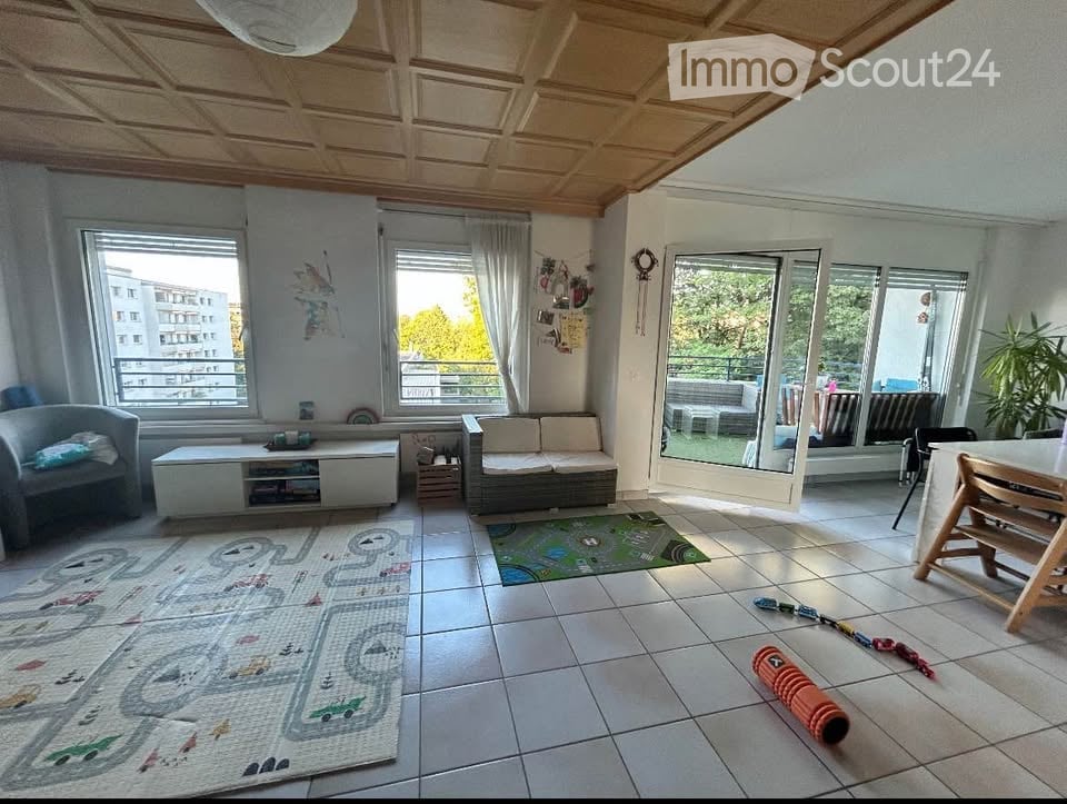 Bright living room with large windows and a sliding glass door leading to a balcony. Features a grey sofa, armchair, coffee table, dining table with chairs, and colorful rugs on the tiled floor.