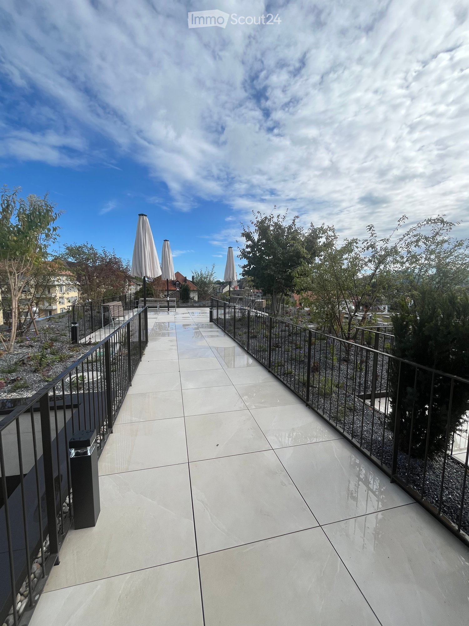 Modern balcony with white tiled flooring, black metal railing, and closed umbrellas in the background.