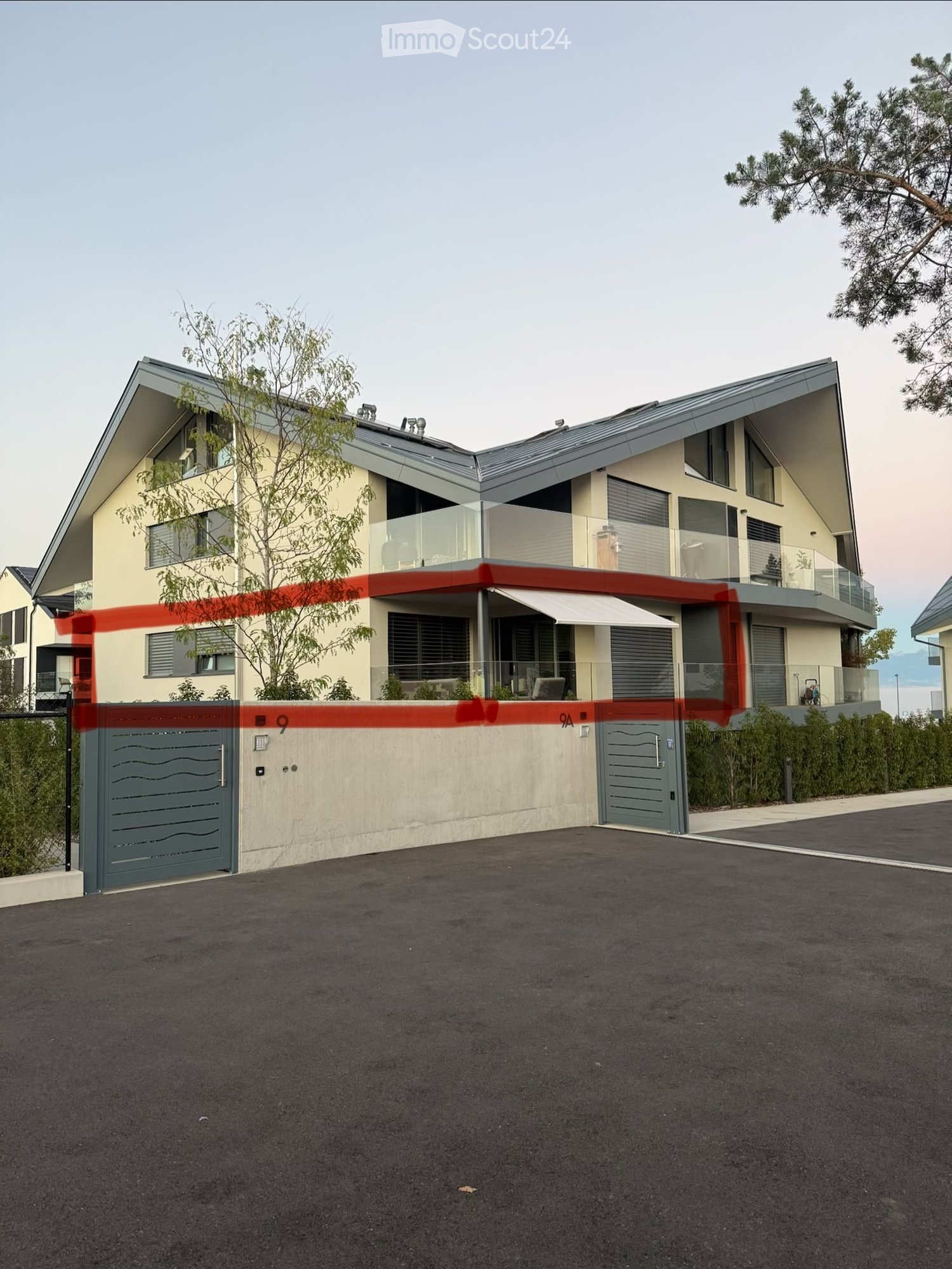 Modern two-story house with a grey roof, large windows, and a balcony area. The house features a contemporary design with a mix of beige and grey exterior walls, and a covered entrance with an awning.