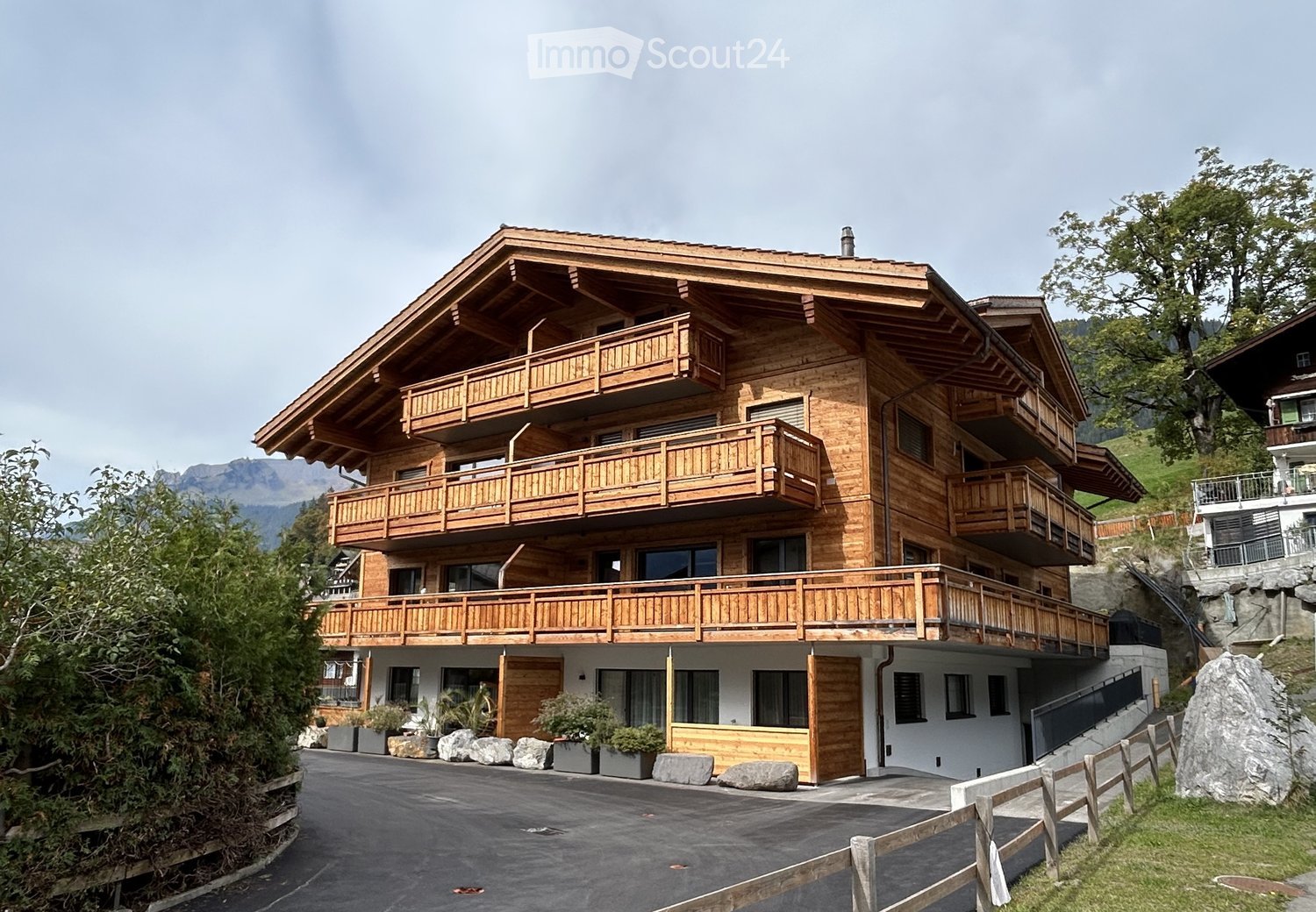 A large, multi-story wooden house with multiple balconies, situated in a scenic mountainous area with a paved driveway and surrounding greenery.