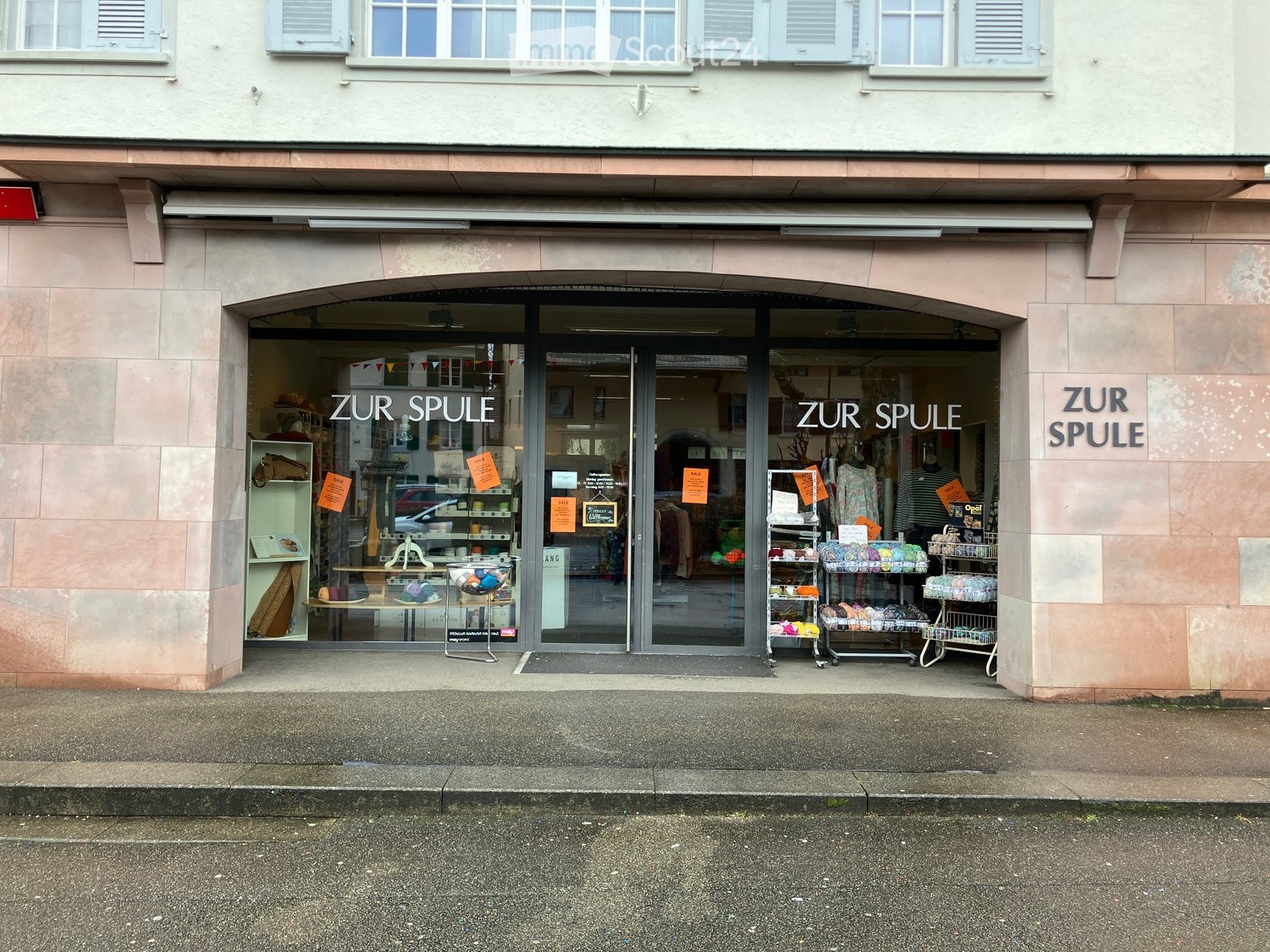 Storefront of a shop with glass doors and windows, name board 'ZUR SPULE', displayed products and banners