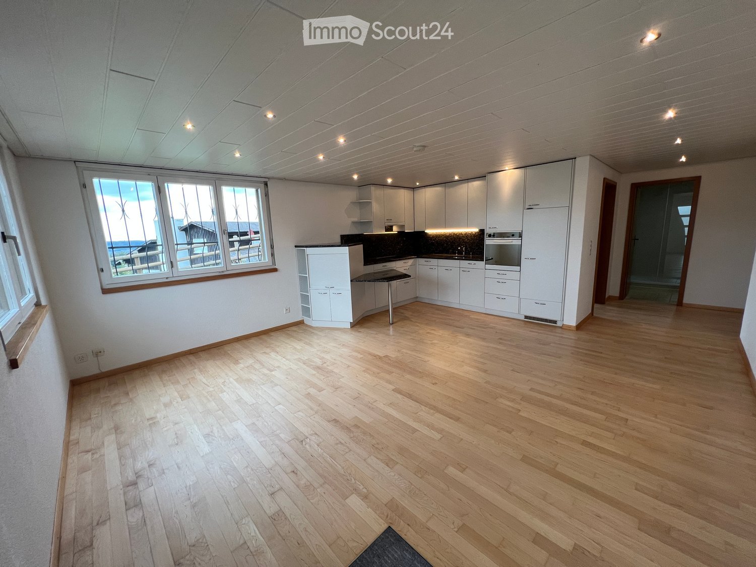 Empty room with light wooden flooring, white walls, and white cabinetry. Large windows provide natural light, and the space is unfurnished.