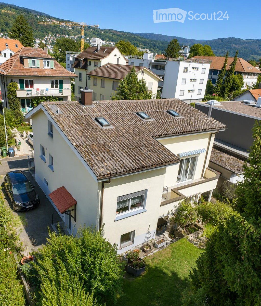 A two-story house with a tiled roof, white exterior, multiple windows, and a small garden area with potted plants.