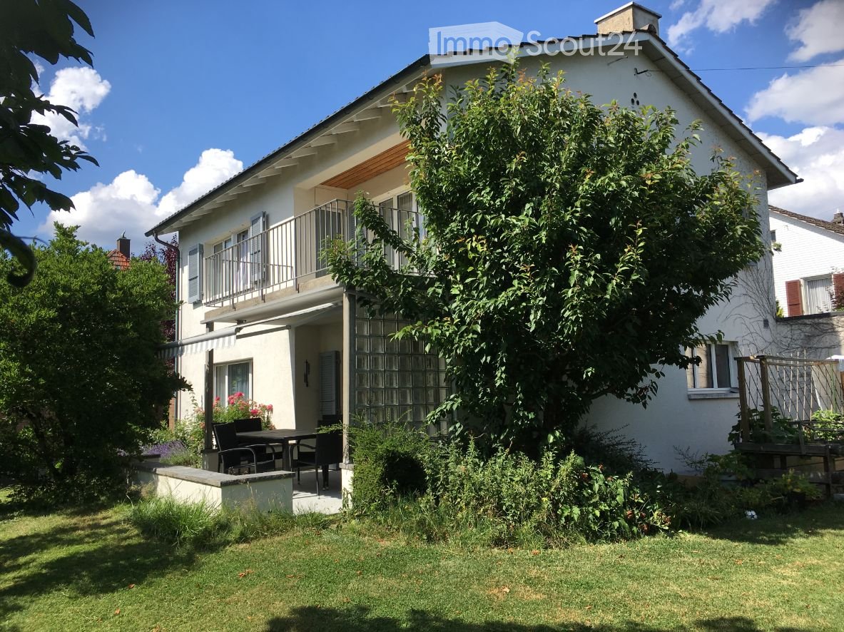 Two-story white house with a balcony, surrounded by a well-maintained garden and lawn.